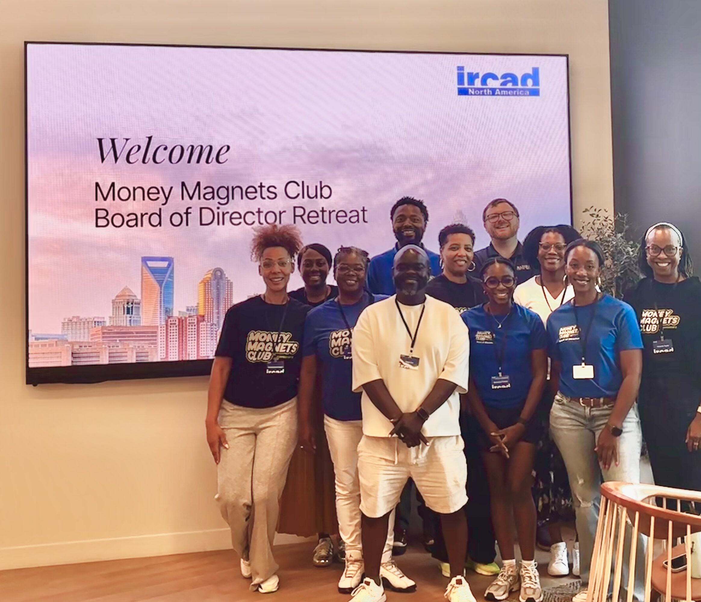 Group of people standing in front of a screen displaying 'Welcome Money Magnets Club Board of Director Retreat' and a logo, smiling.