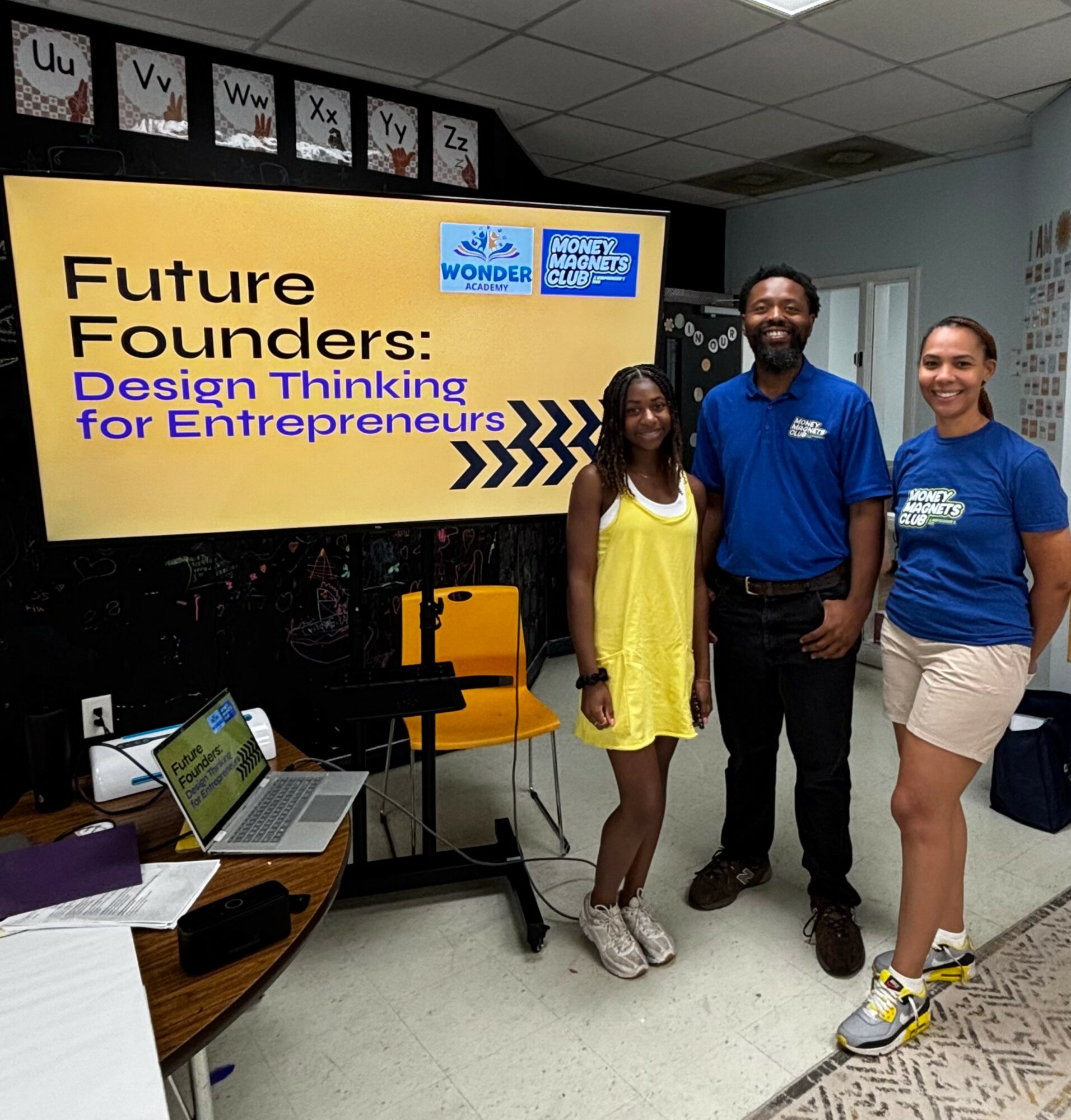 Three people standing in front of a large screen with a presentation slide, smiling, in a classroom setting.