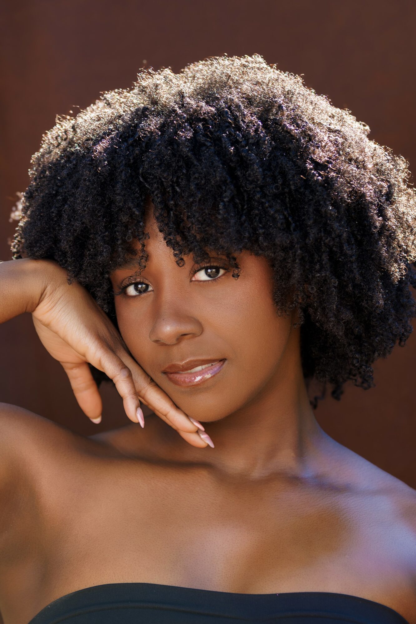 Portrait of a woman with natural curly hair, resting her chin on her hand, looking at the camera.