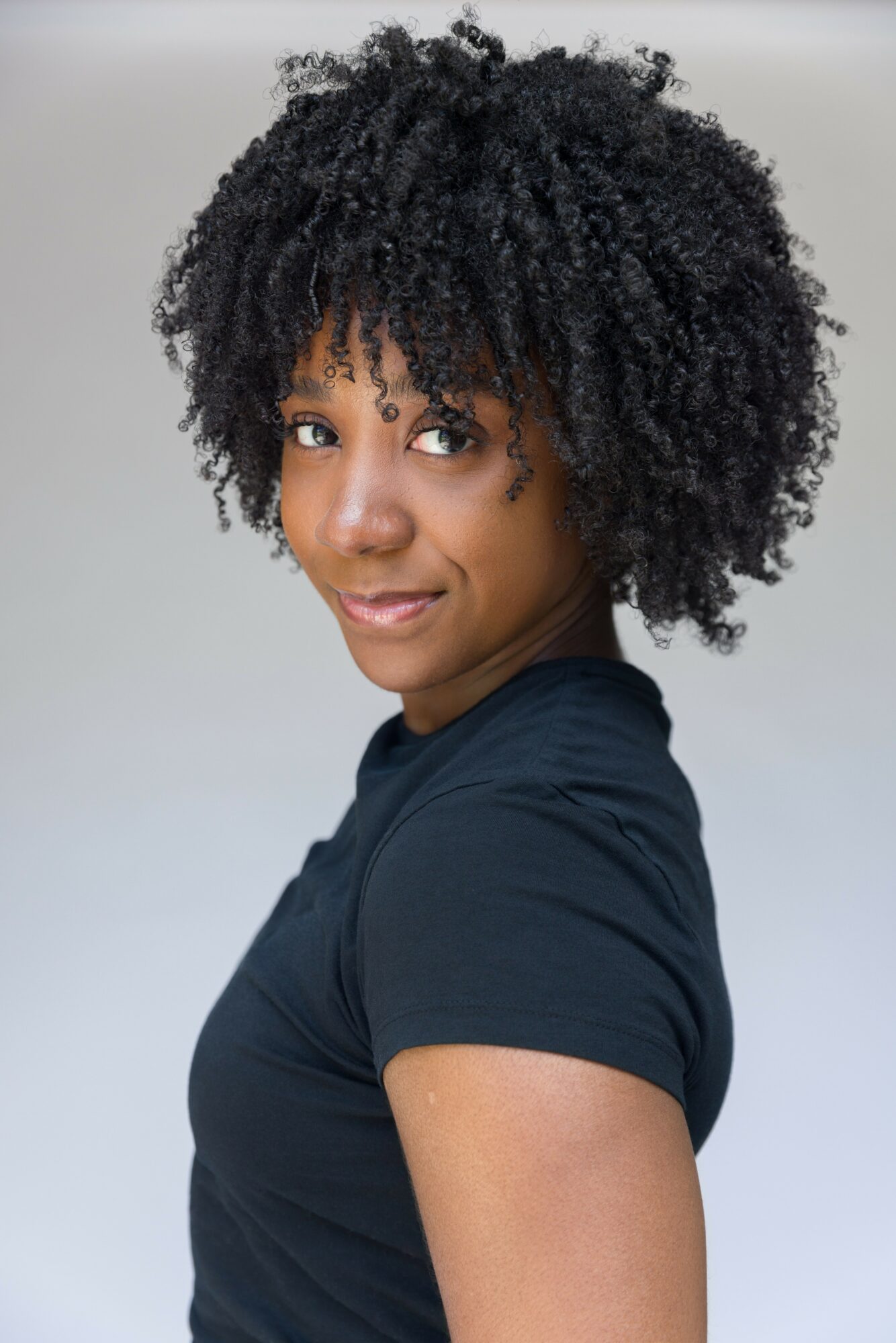 Woman with curly hair wearing a black shirt, looking at the camera, smiling slightly, against a plain background.