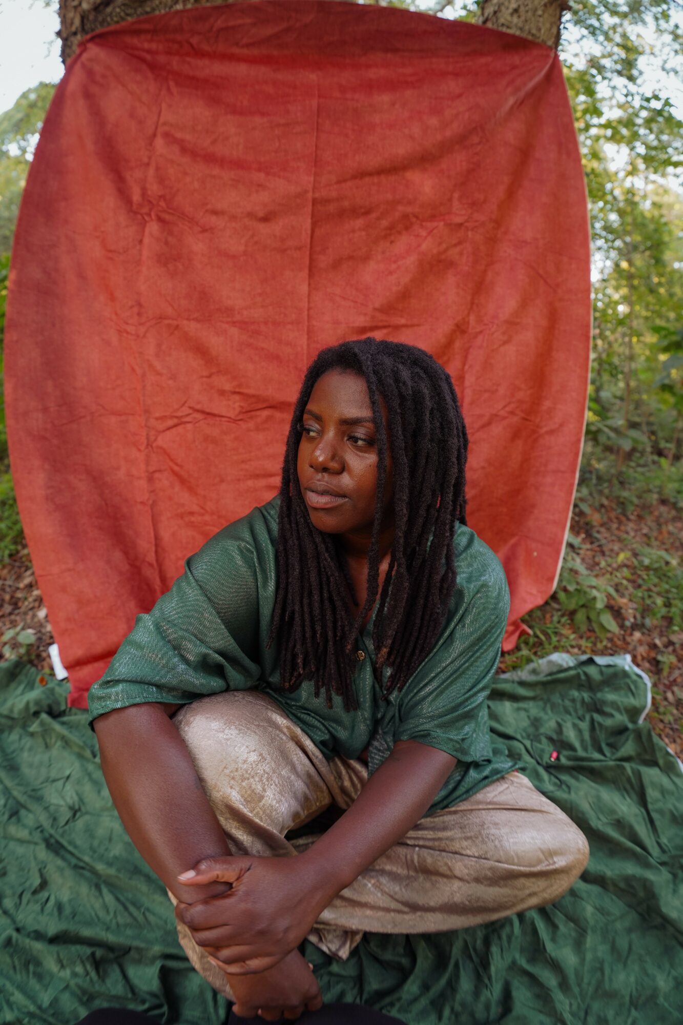 Person with long dreadlocks sitting outdoors in front of a red backdrop, wearing a green shirt and beige pants.