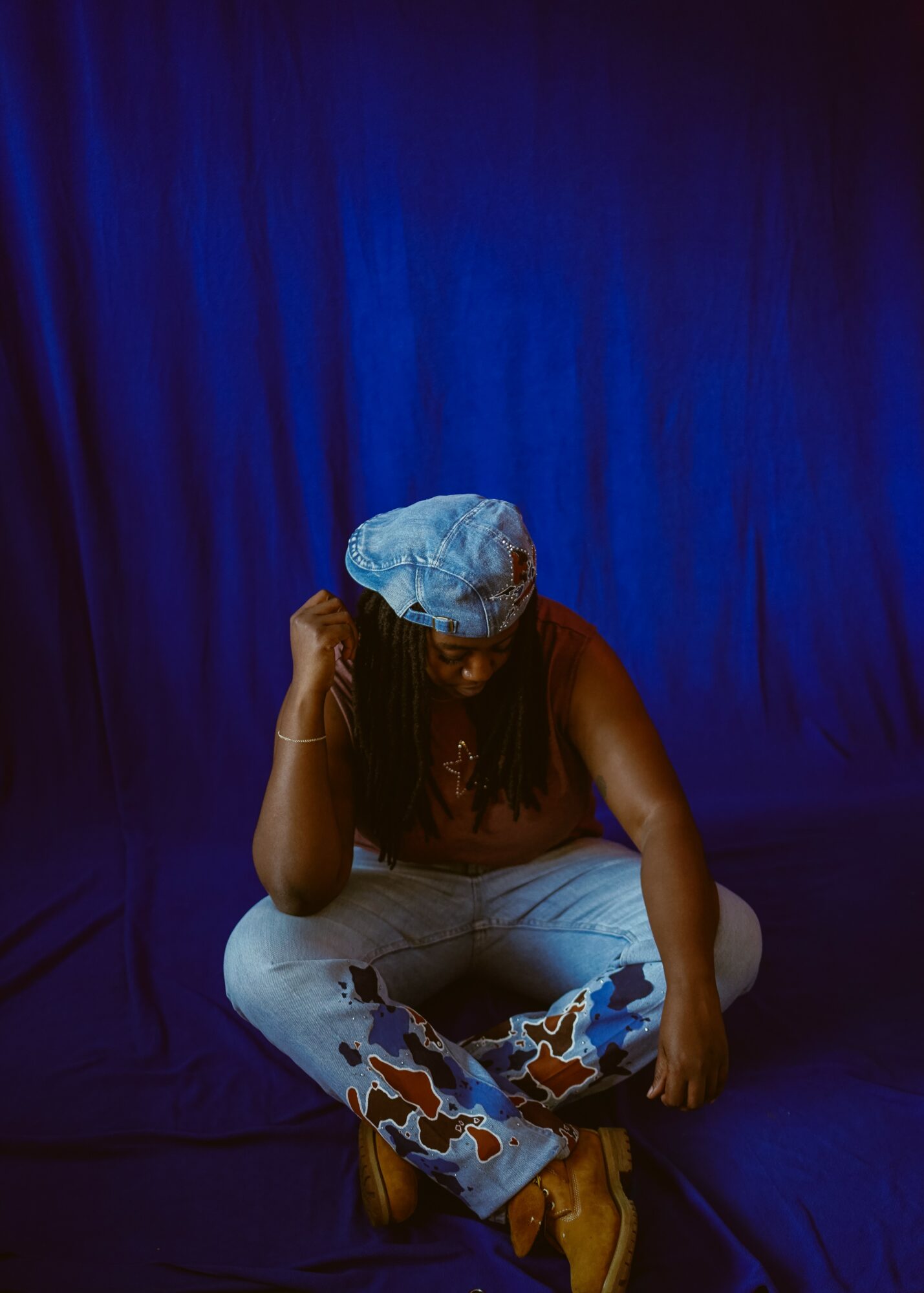 Person sitting cross-legged on stage with a blue curtain background, wearing a blue head covering and casual clothing.