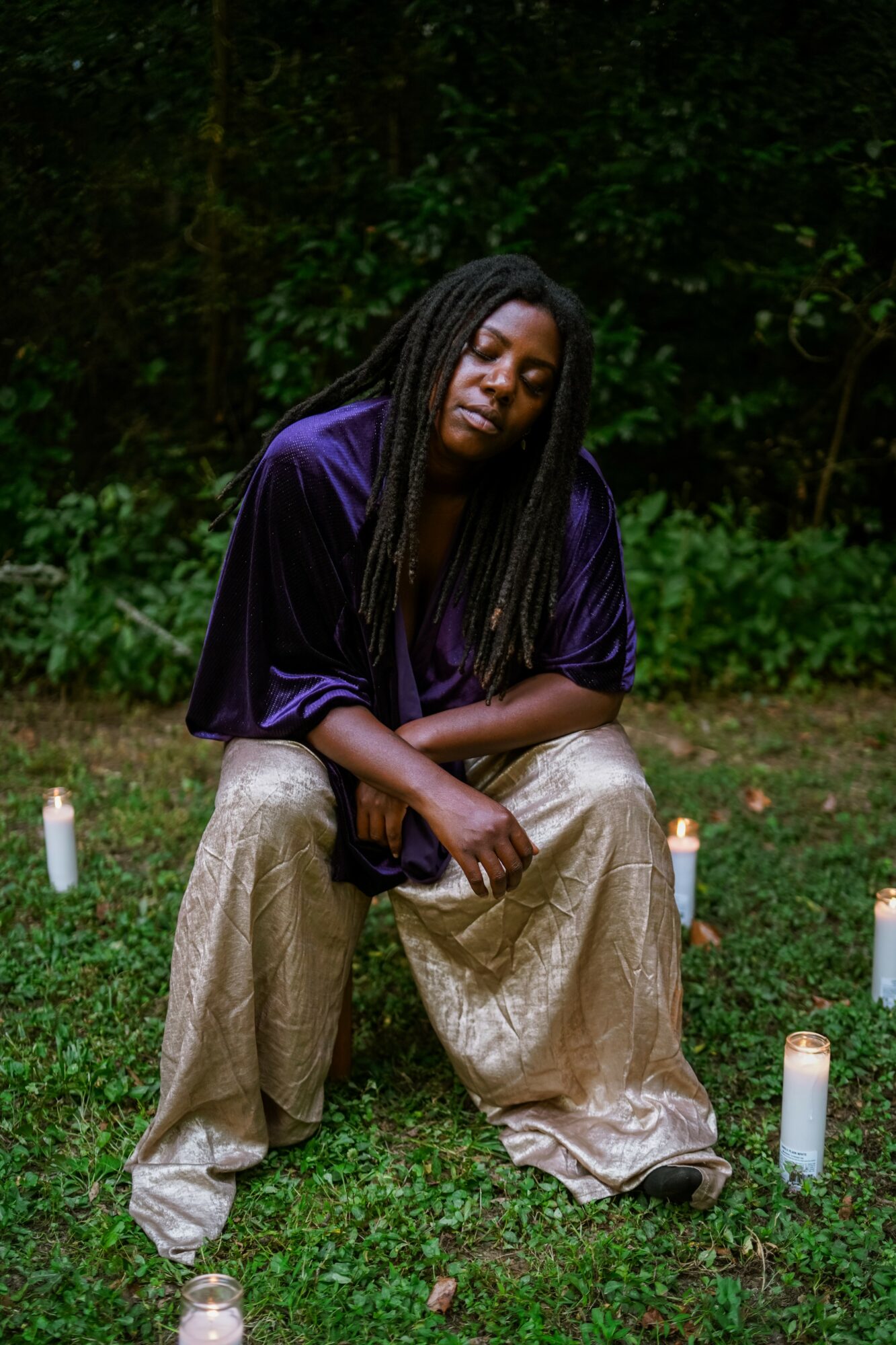Person with long braided hair sitting outdoors on grass, surrounded by candles, with a dark green background.