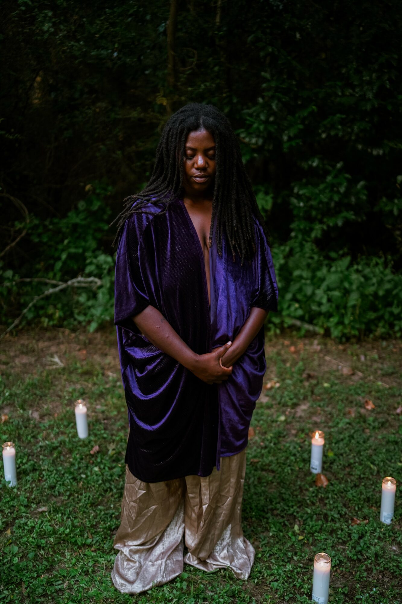 A woman with long dreadlocks stands outdoors in a forested area, wearing a purple velvet top and beige pants, surrounded by candles.