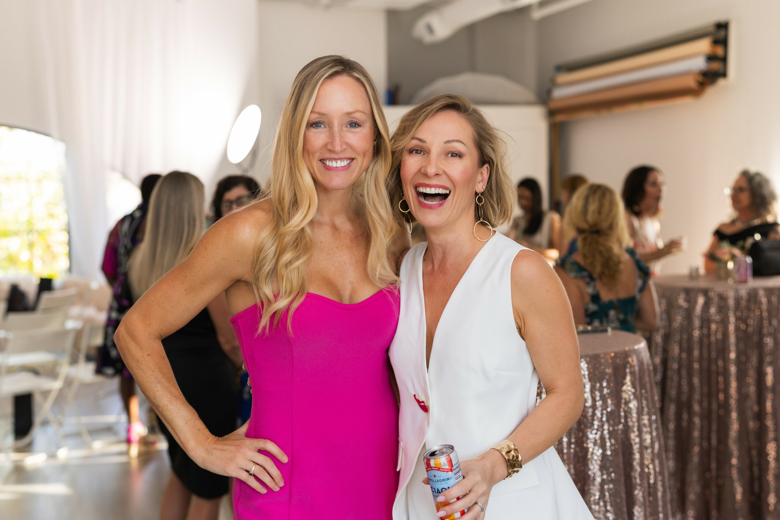 Two women smiling and laughing at a social event, with other people in the background, indoors.