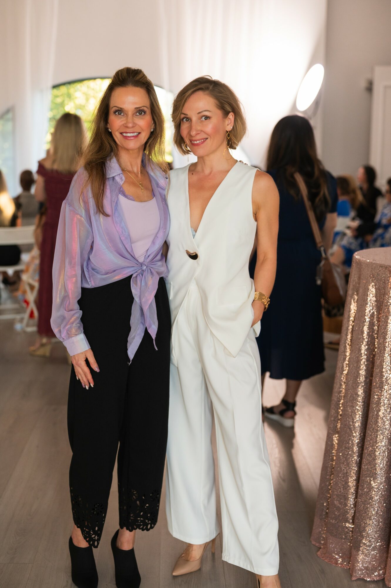 Two women stand together indoors, smiling, with people and tables in the background, one in a purple blouse and black pants, the other in a white jumpsuit.