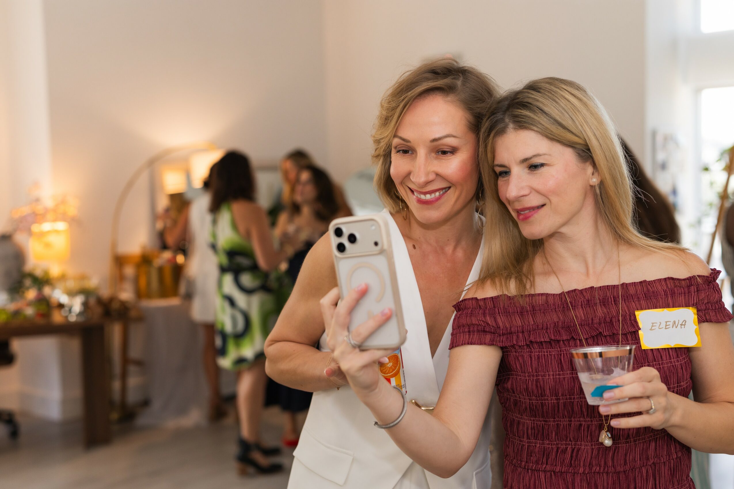Two women smiling and looking at a smartphone at a social gathering, one holding a drink, in a well-lit room.