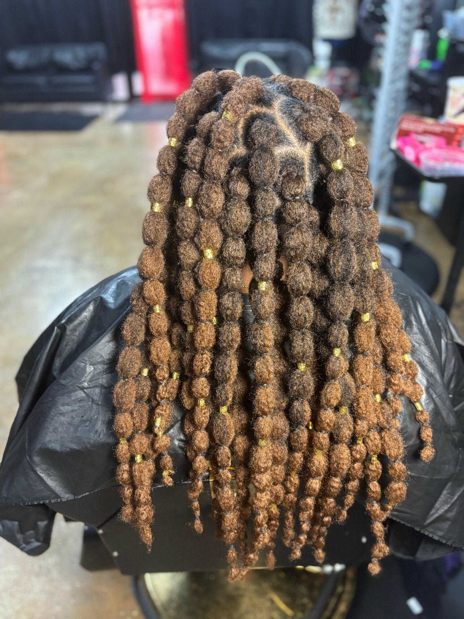 Person with long, thick, brown dreadlocks decorated with small gold beads, sitting in a salon chair.