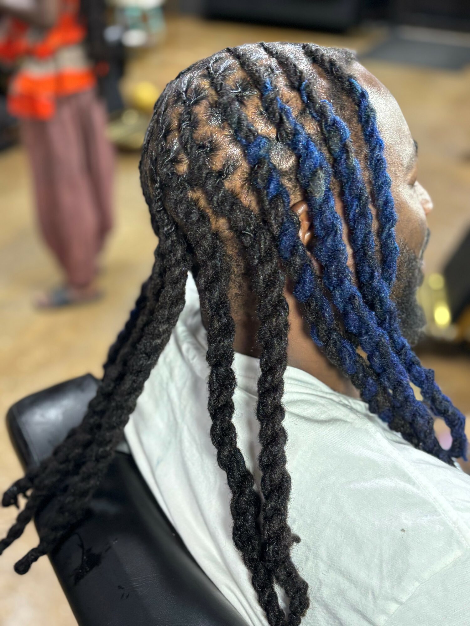 Side view of a person with styled dreadlocks, some dyed blue, wearing a white cape, in a salon setting.