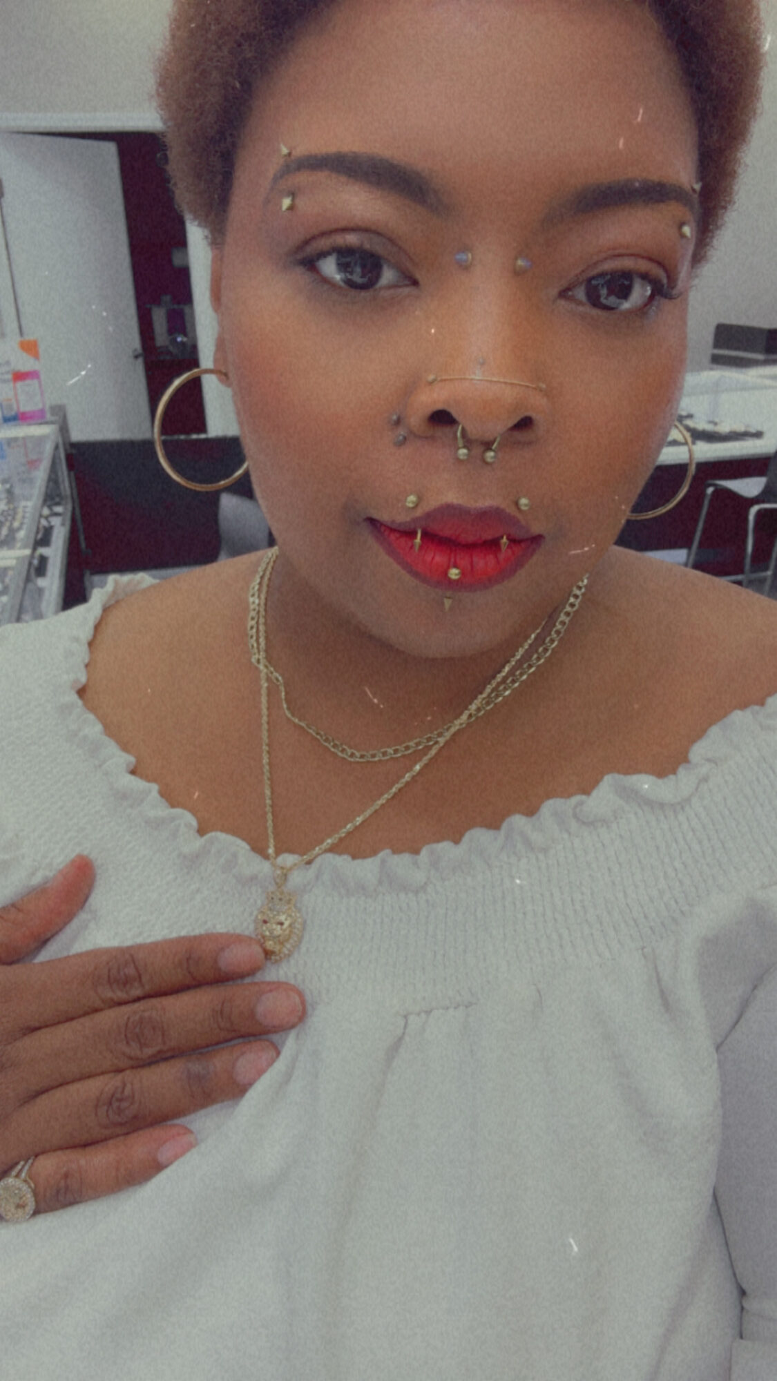 Close-up of a person with short hair, multiple facial piercings, red lipstick, wearing a white top and gold jewelry.