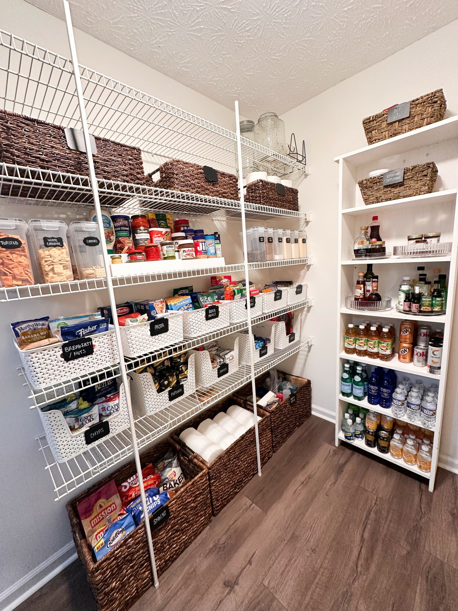 Pantry with white wire shelves filled with baskets, jars, and food items, and a white shelf with bottles and jars, against a beige wall.