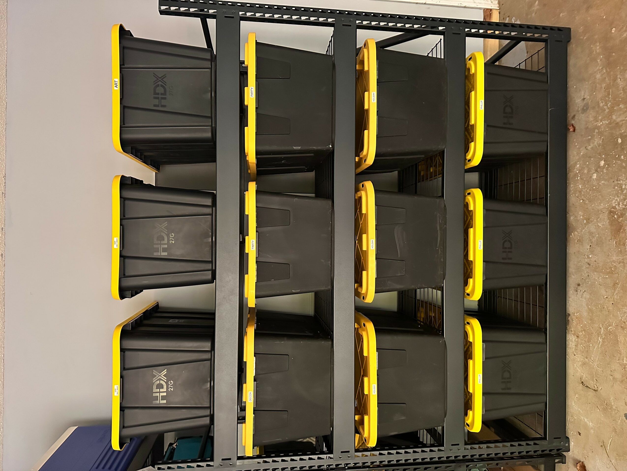 Black storage bins with yellow handles on a metal shelving unit against a white wall.