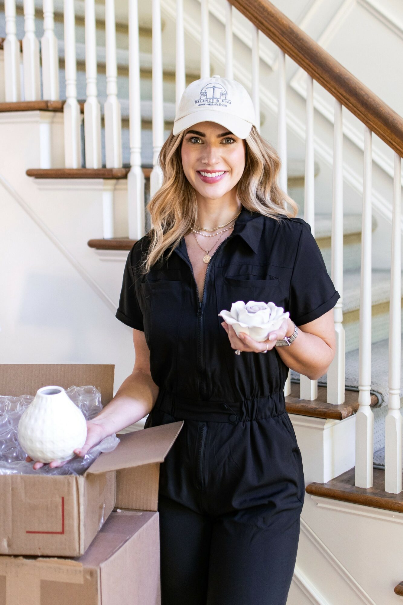 Woman in black outfit holding a cupcake, standing near staircase, smiling, wearing a white cap, with boxes and a teapot nearby.