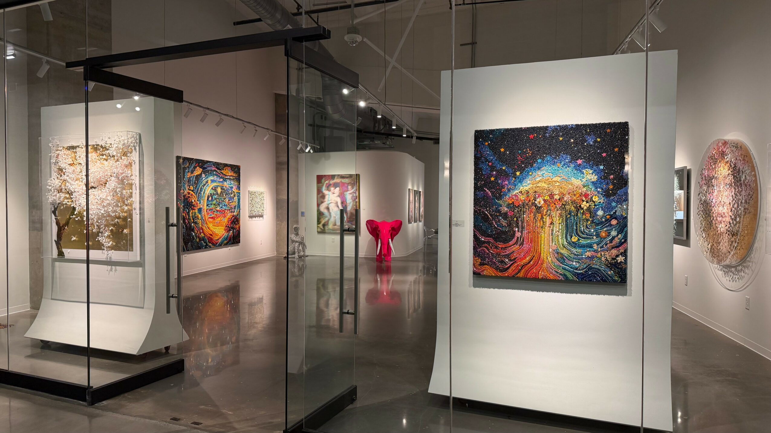 Gallery with colorful abstract paintings on white walls and a glass display case, reflecting overhead lighting.