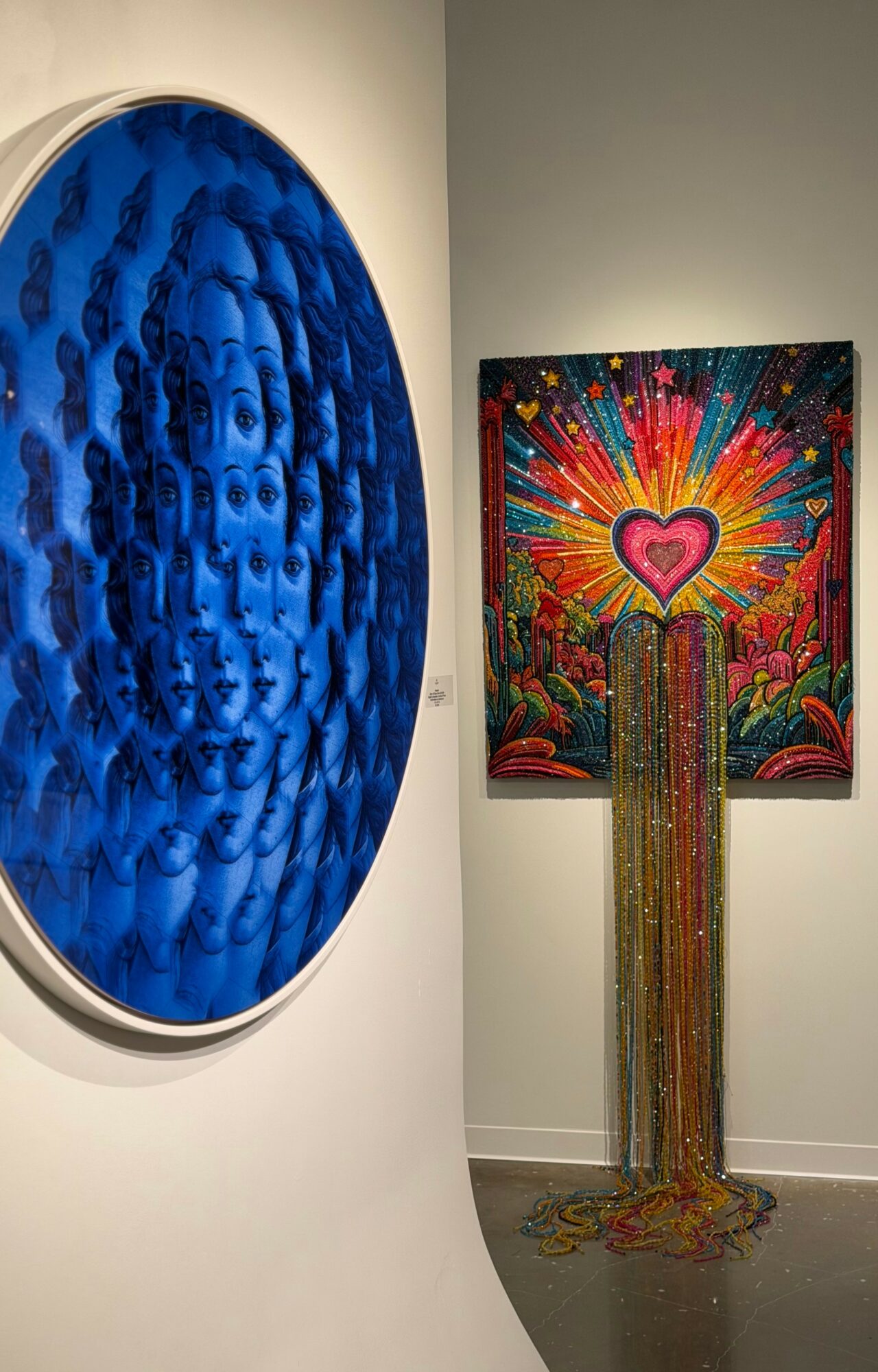 Two colorful artworks displayed on gallery walls, one circular blue piece and one rectangular with a heart and rainbow colors, with beads hanging below.