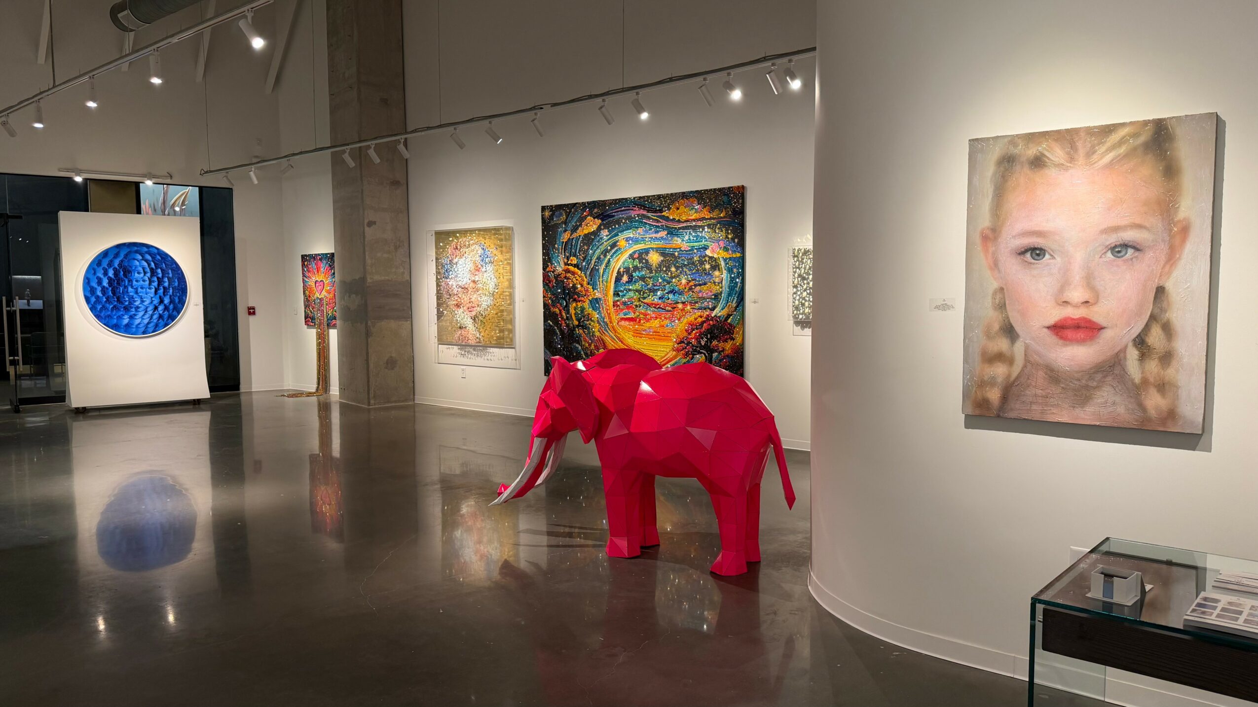 Pink elephant sculpture in an art gallery with paintings on the walls, including a portrait of a girl with braided hair.