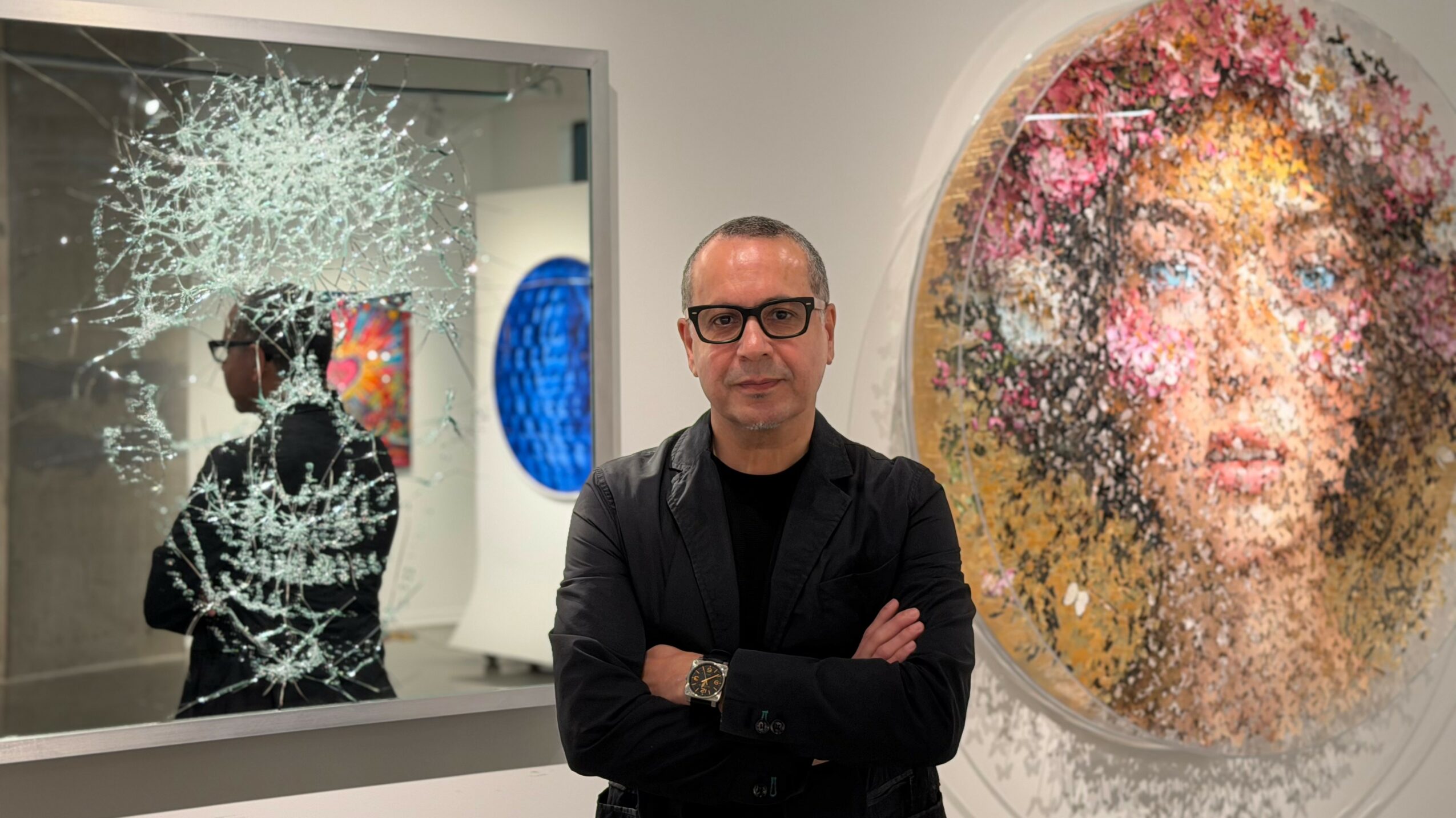 Man with glasses and black jacket standing with arms crossed in art gallery, colorful artwork behind him.