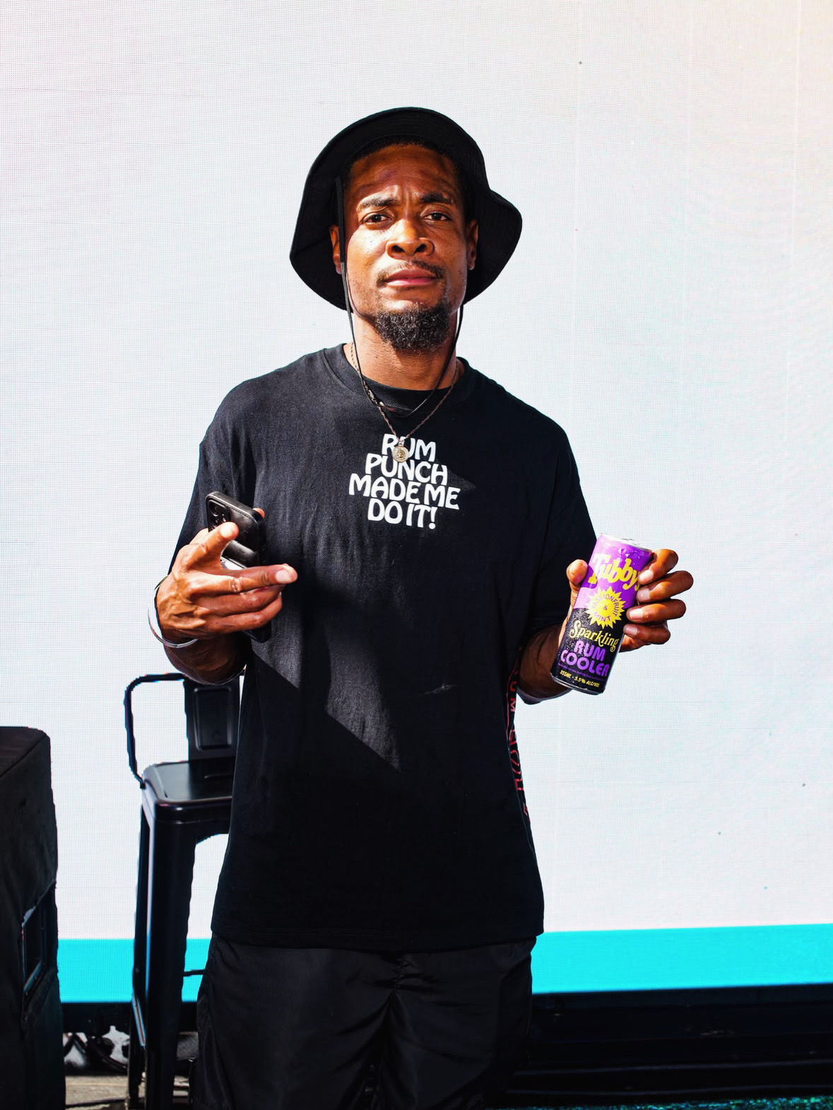 Man wearing a black hat and T-shirt holding a phone in one hand and a colorful drink in the other.