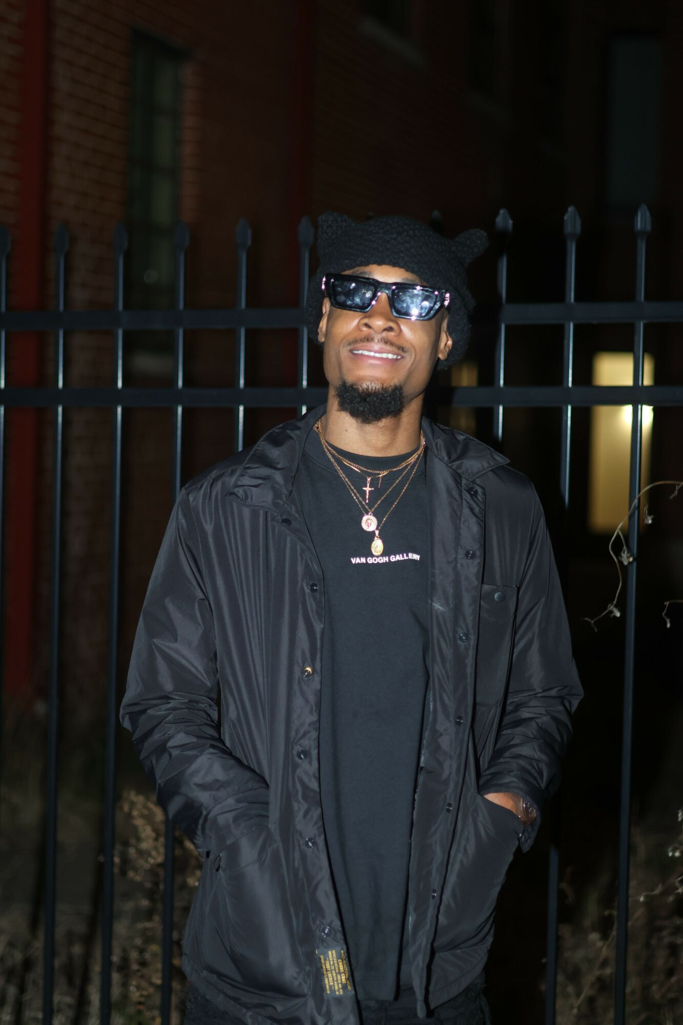 Man wearing sunglasses and a black jacket, smiling outdoors at night, with a metal fence behind him.