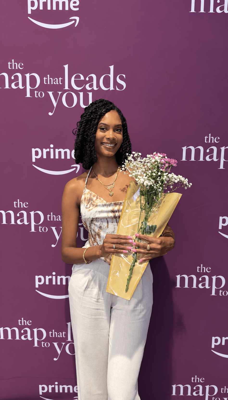 Young woman with long dark hair holding a bouquet of flowers and a paper bag, smiling, standing in front of a purple backdrop.