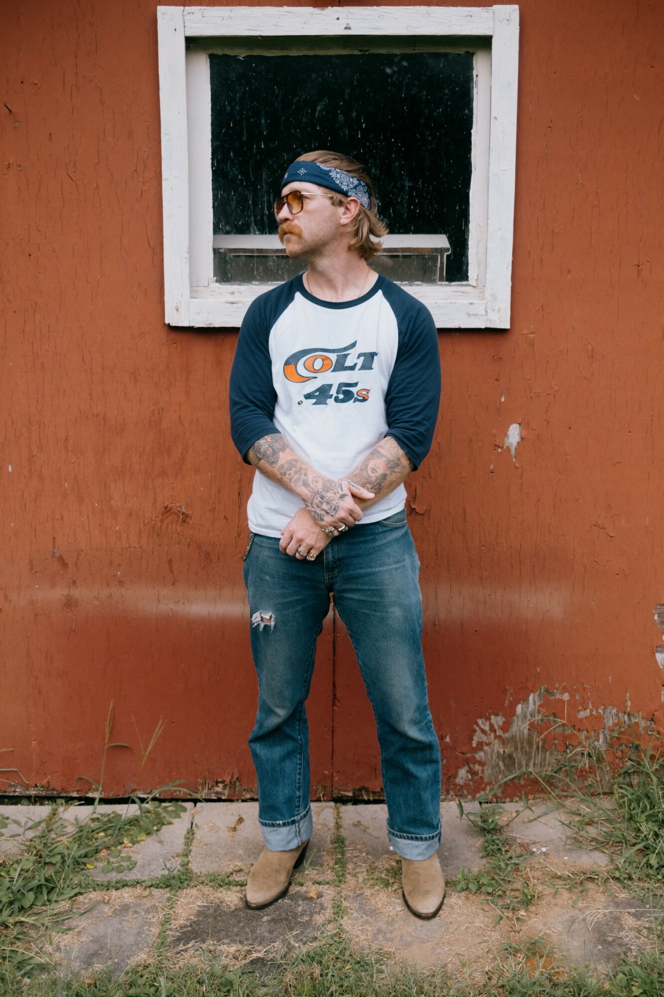 Man with tattoos and a headband stands outdoors against an orange wall with a window, looking to the side.