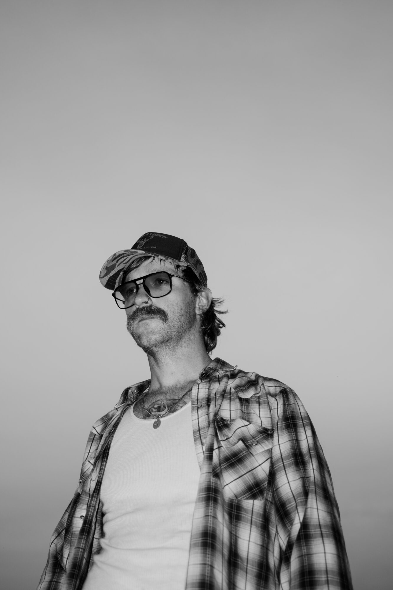 Man with glasses, beard, and mustache wearing a plaid shirt and cap, looking to the side, against a plain background.