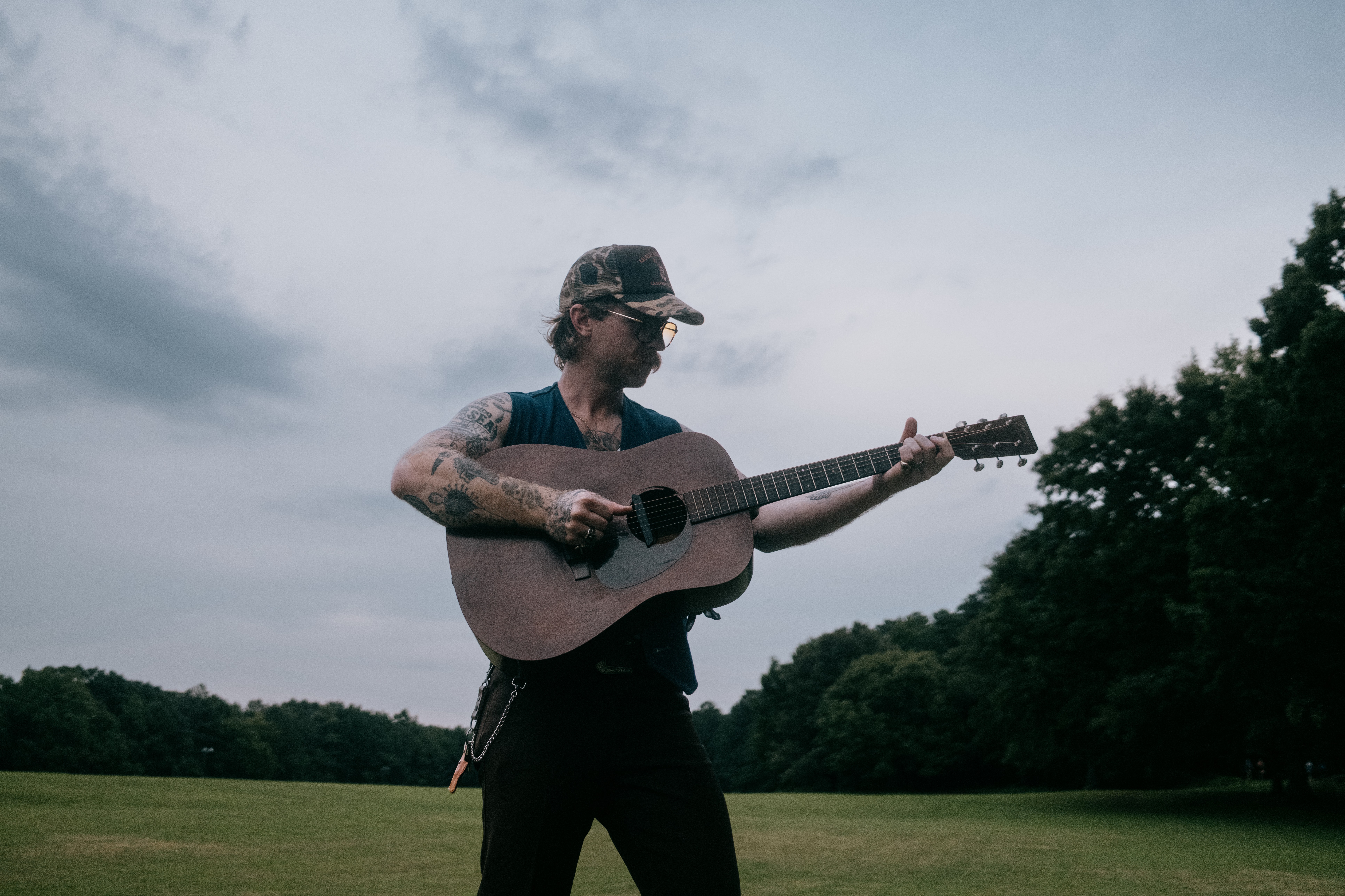 Person playing guitar outdoors with trees and cloudy sky in background.