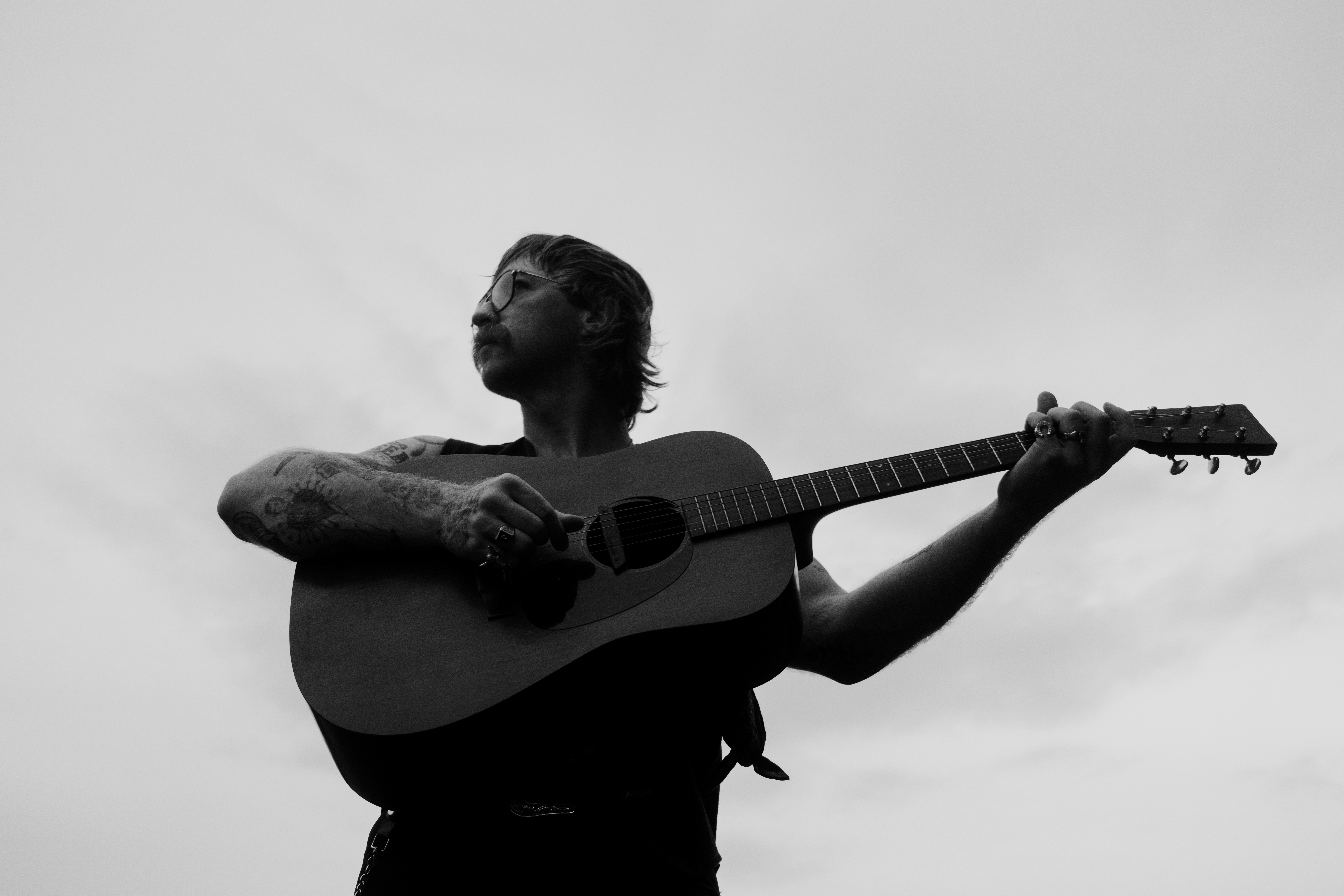 Person playing guitar, looking upward, with arm tattoos, in black and white.
