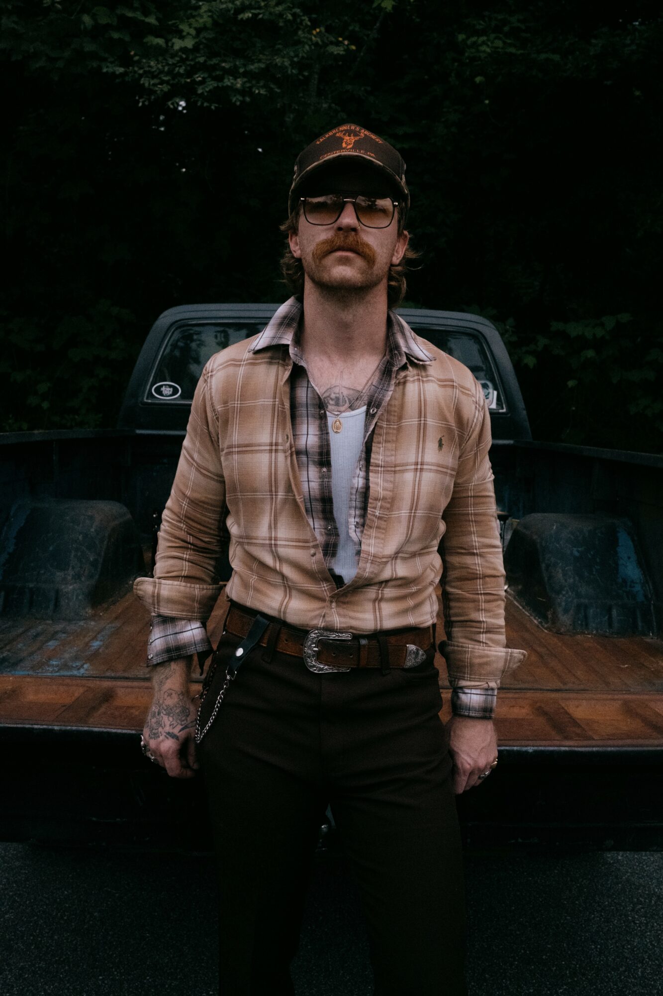 Man with sunglasses and tattoos stands in front of a truck, wearing a plaid shirt and cap, outdoors at night.