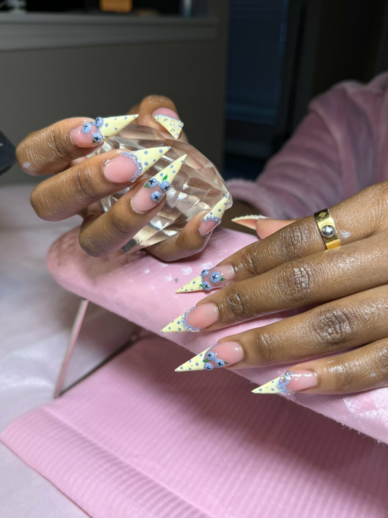 Person with decorated nails holding a clear container, wearing a pink garment and a gold ring, with a pink surface below.