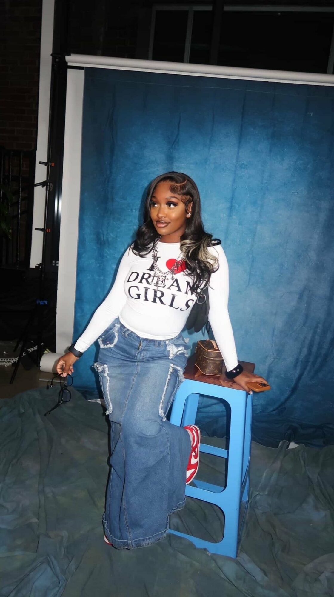Woman with long hair in a white shirt and jeans, standing next to a blue stool against a blue backdrop.