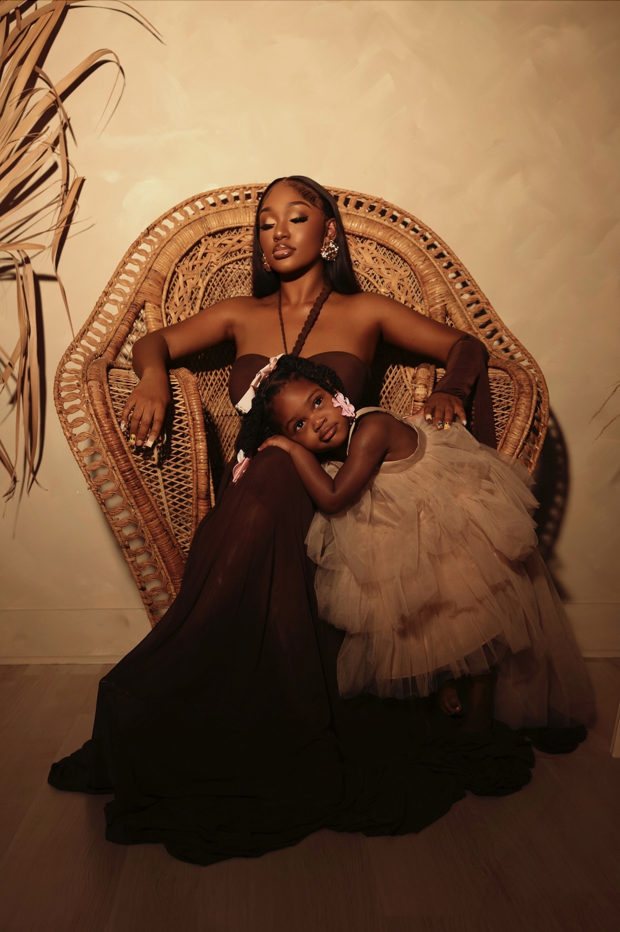 Two women pose on a wicker chair, one sitting and one leaning on her, in a warm-toned room.