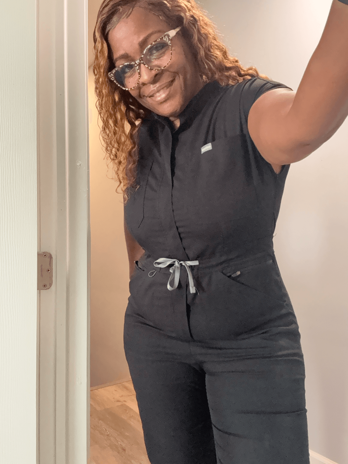 Woman with curly hair and glasses taking a selfie, smiling, wearing a black outfit, in a room with a door.