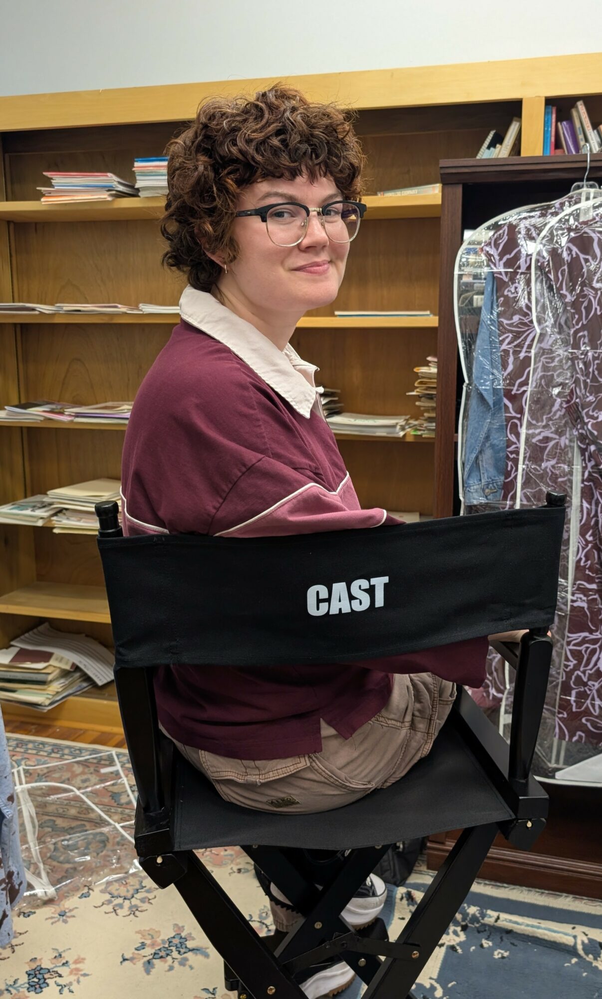 Person with curly hair and glasses sitting in a chair in a room with bookshelves, wearing a maroon sweater and beige pants.