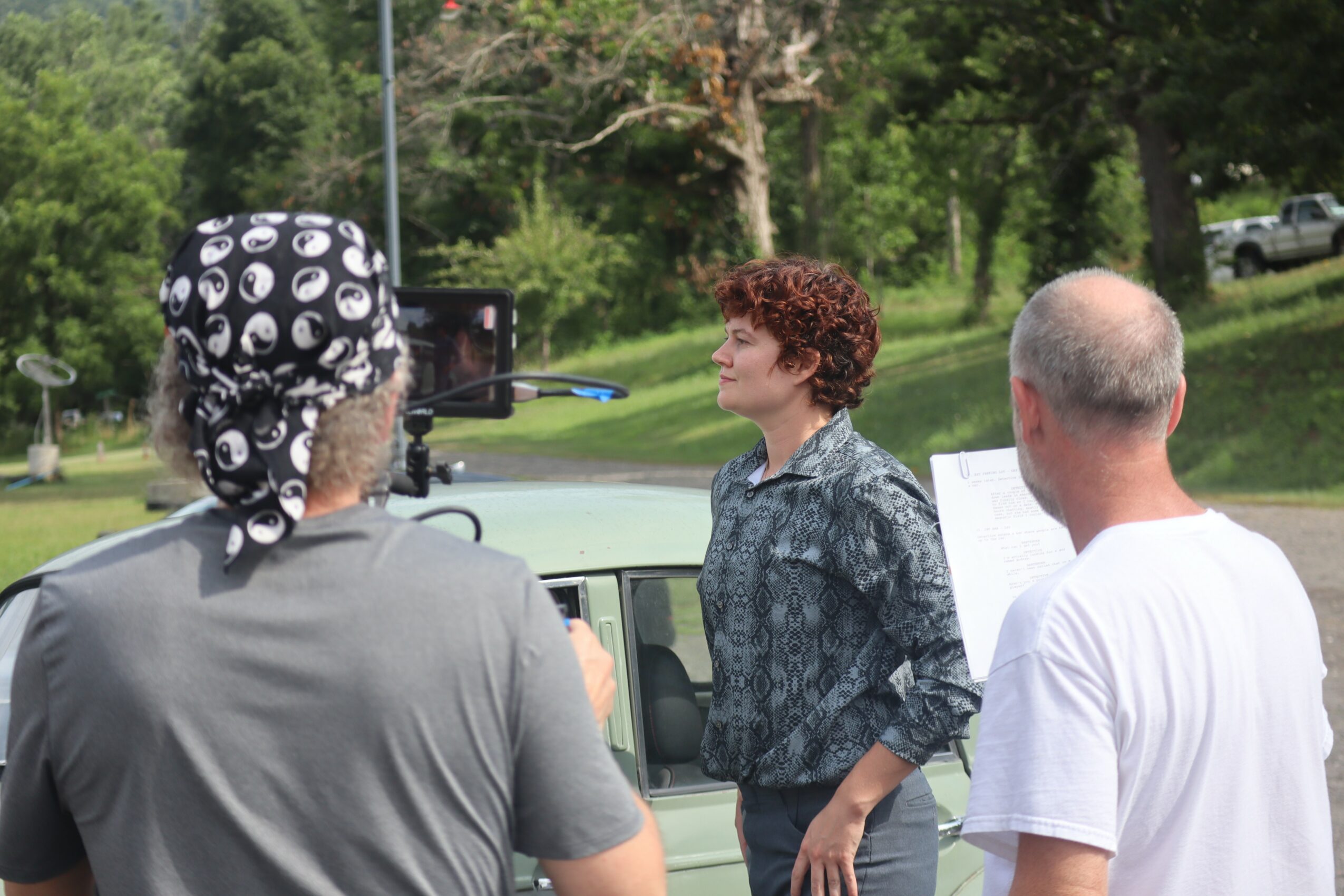 Three people outdoors, one woman with curly hair and two men, one with a bandana, engaged in conversation near a vehicle.
