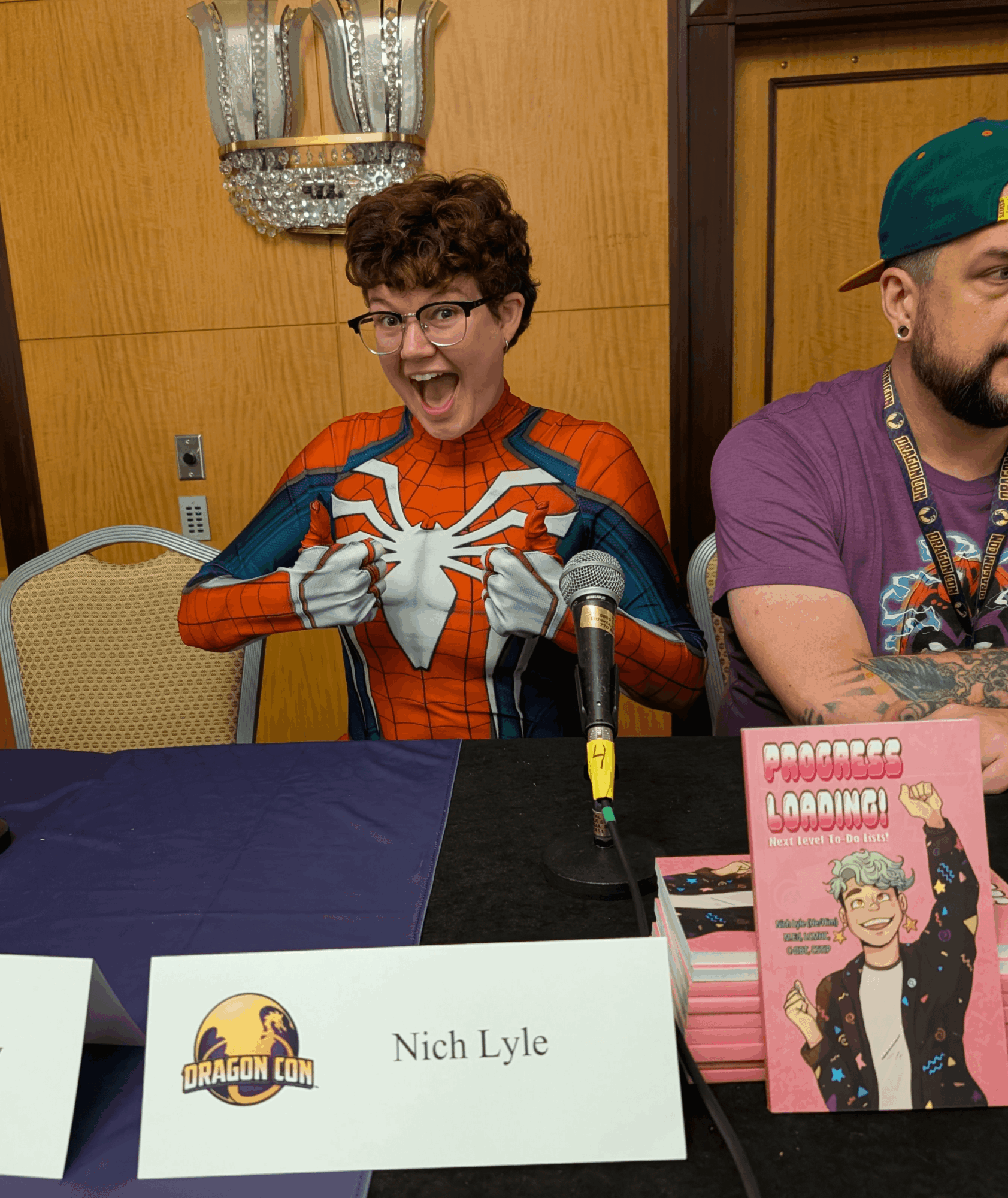 Person dressed as Spider-Man smiling and making a heart shape with hands at a panel event, with a nameplate reading 'Nich Lyle'.