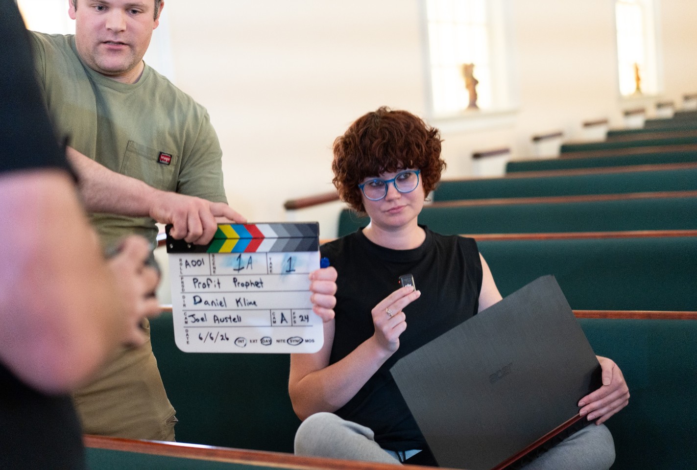 Person with curly hair and glasses sitting in a room with empty seats, holding a laptop and a small object, with a crew member holding a clapperboard nearby.