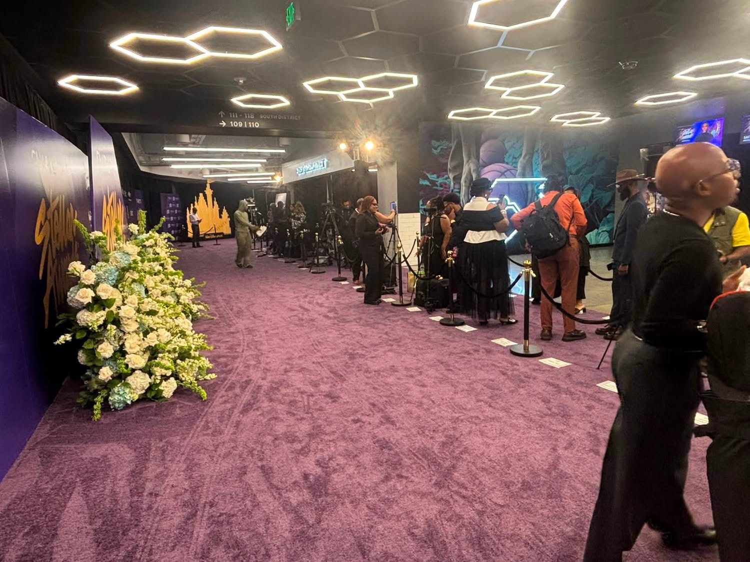 People standing in line at an indoor event with purple carpet and modern ceiling lights, floral arrangement on the left.