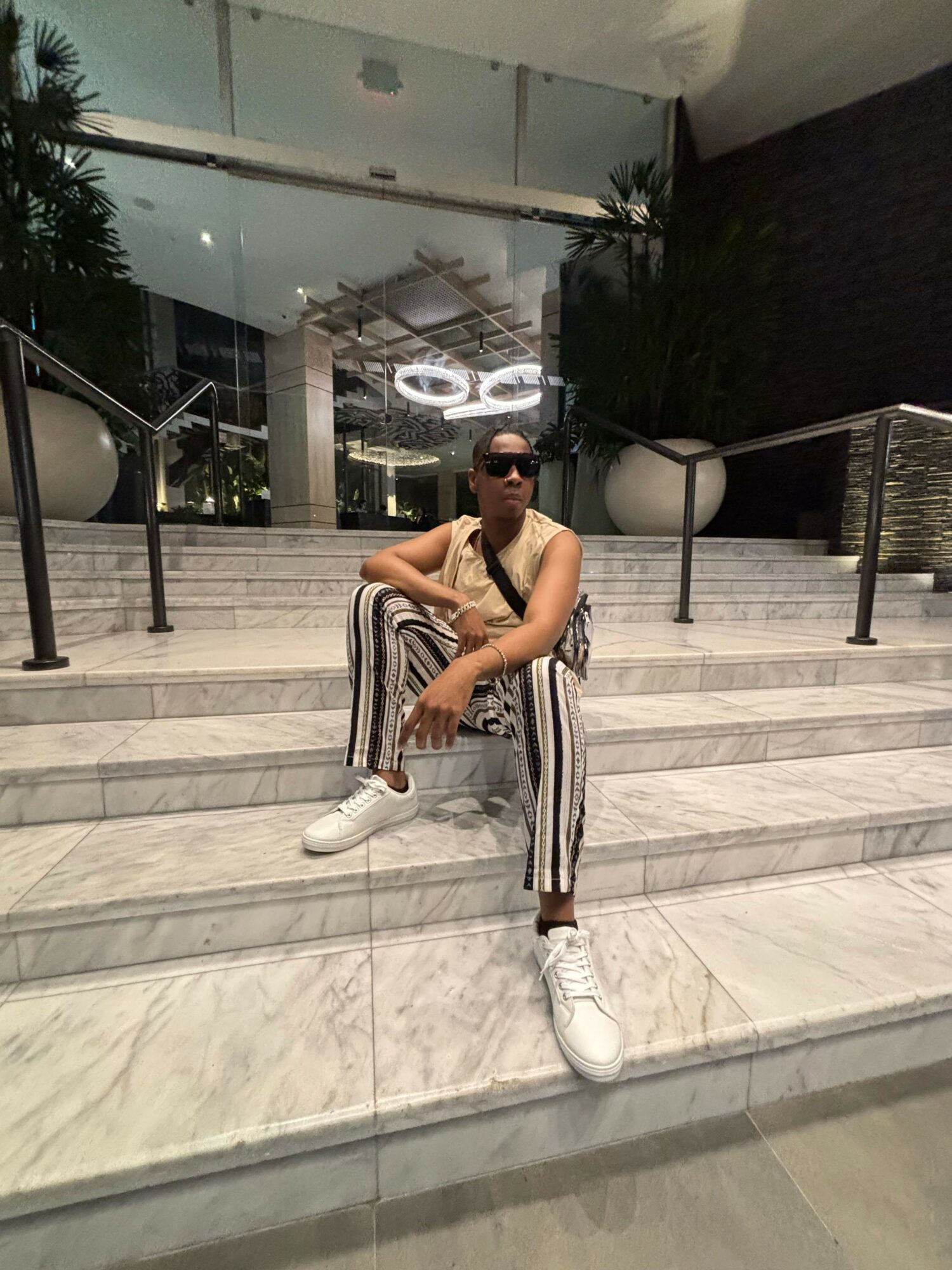 Person crouching on marble stairs outside building, wearing striped pants and white shoes, with glass door and plants in background.