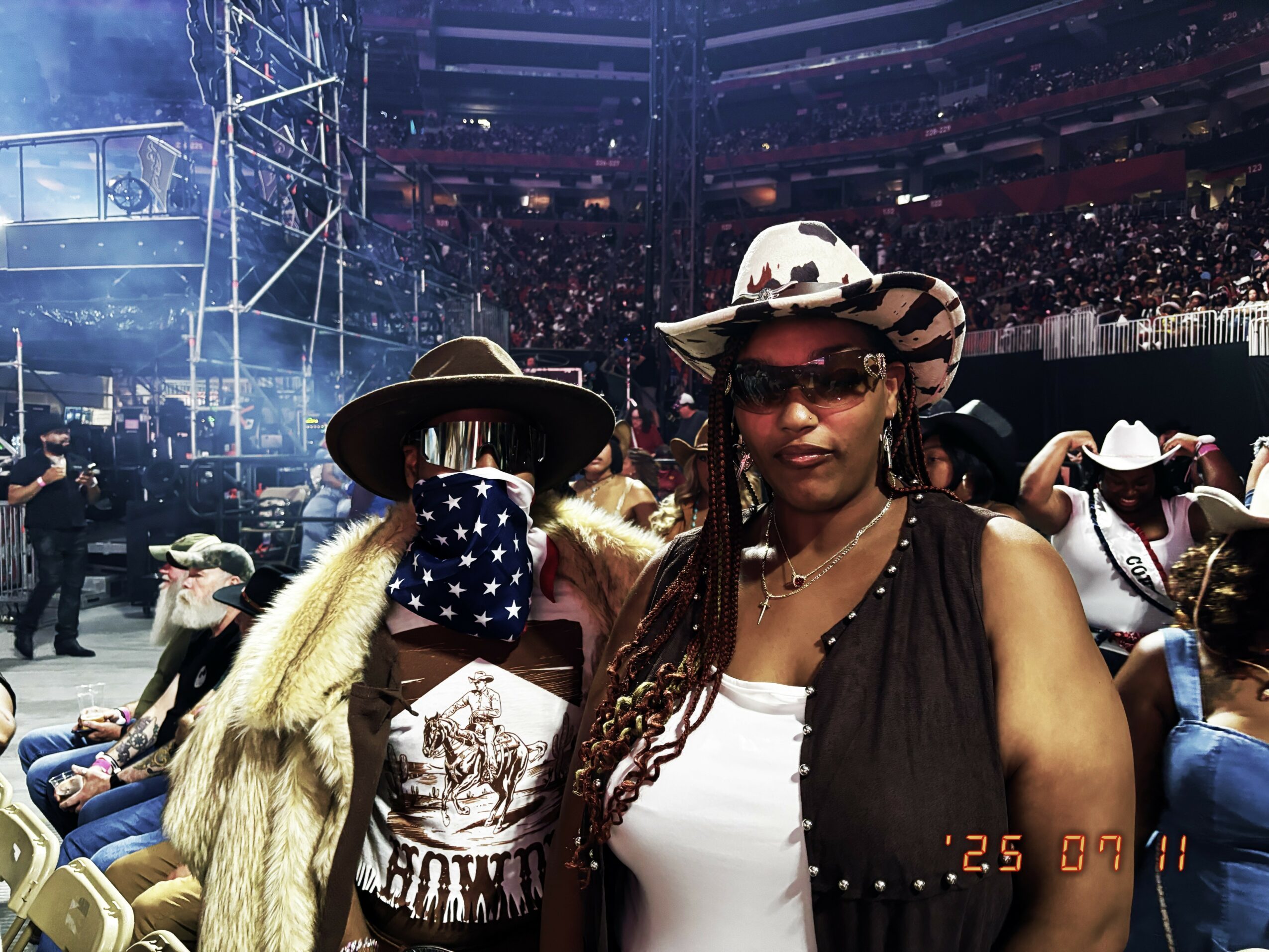 Two women wearing hats and sunglasses at an outdoor event, with a crowd and stage in the background.