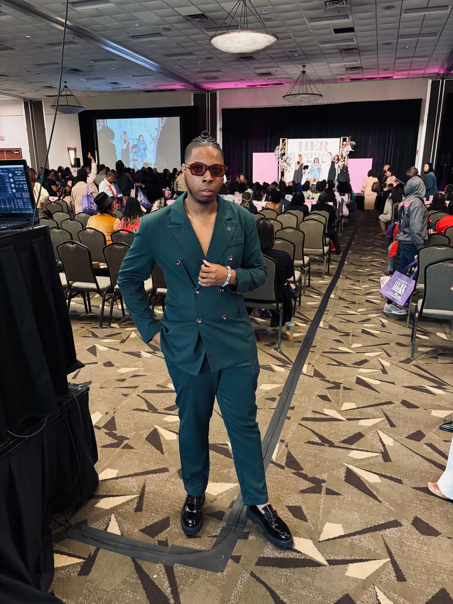 Man in teal suit and sunglasses standing in conference room with audience and stage in background.
