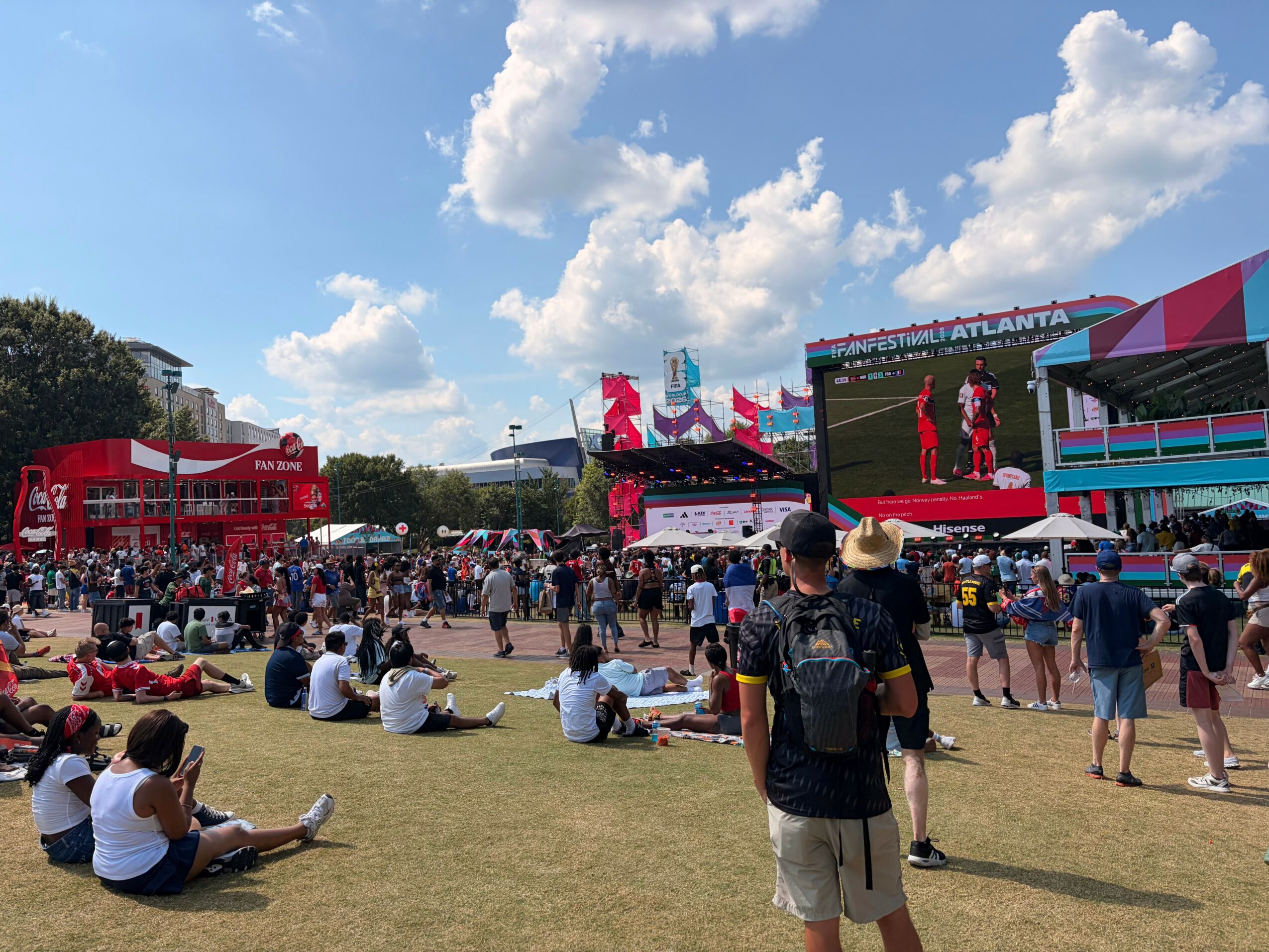 Crowd gathered at outdoor music festival with stage, tents, and blue sky with clouds.