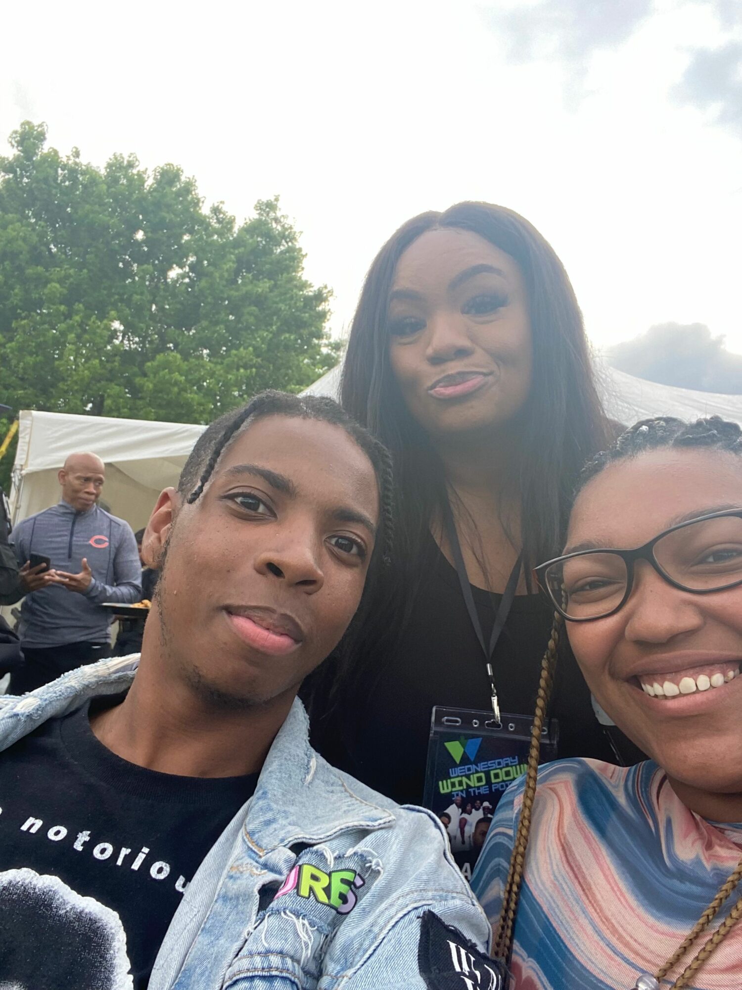 Three people smiling outdoors, with trees and a tent in the background.