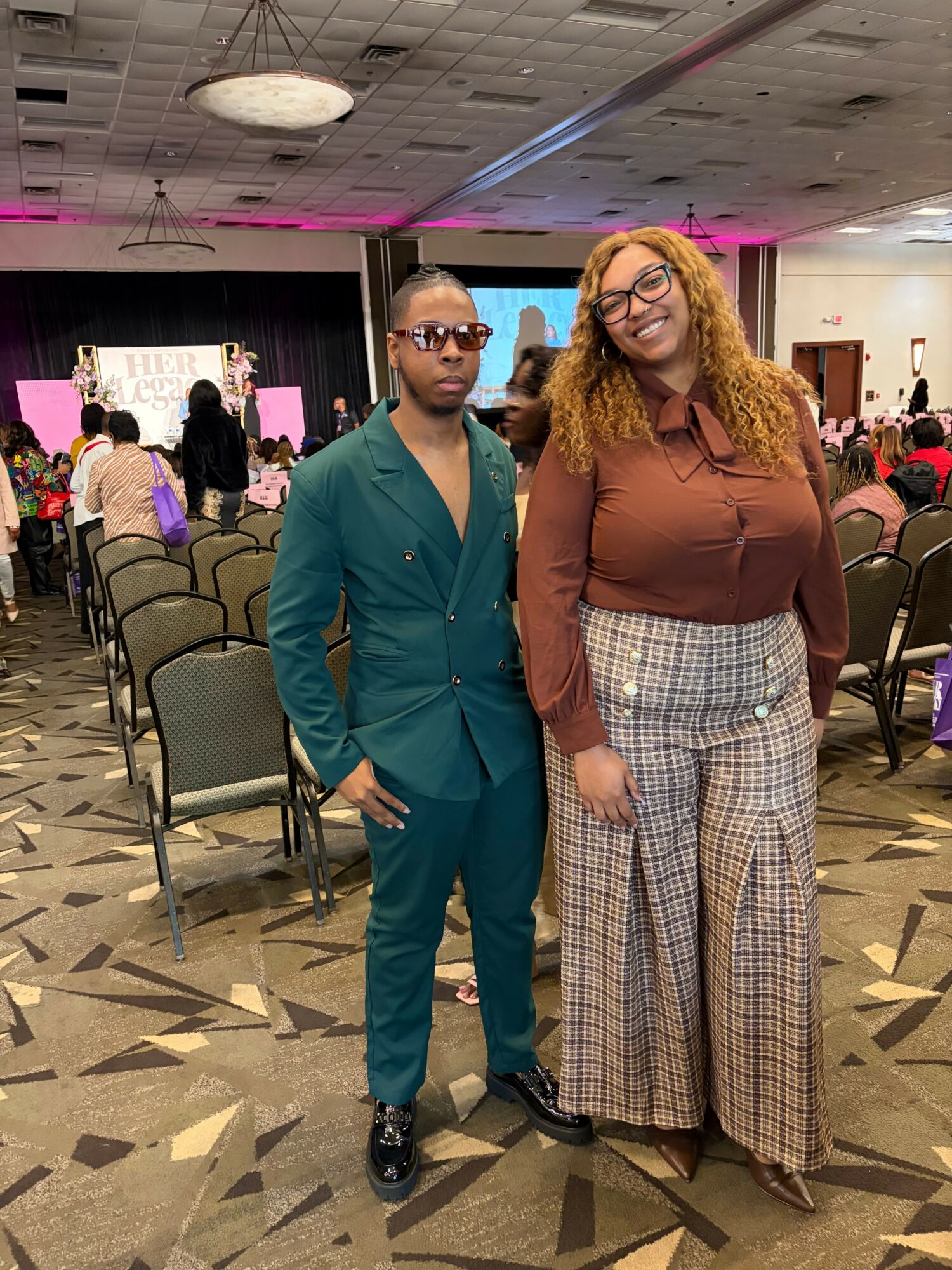 Two people standing in a conference room with chairs and a stage in the background.