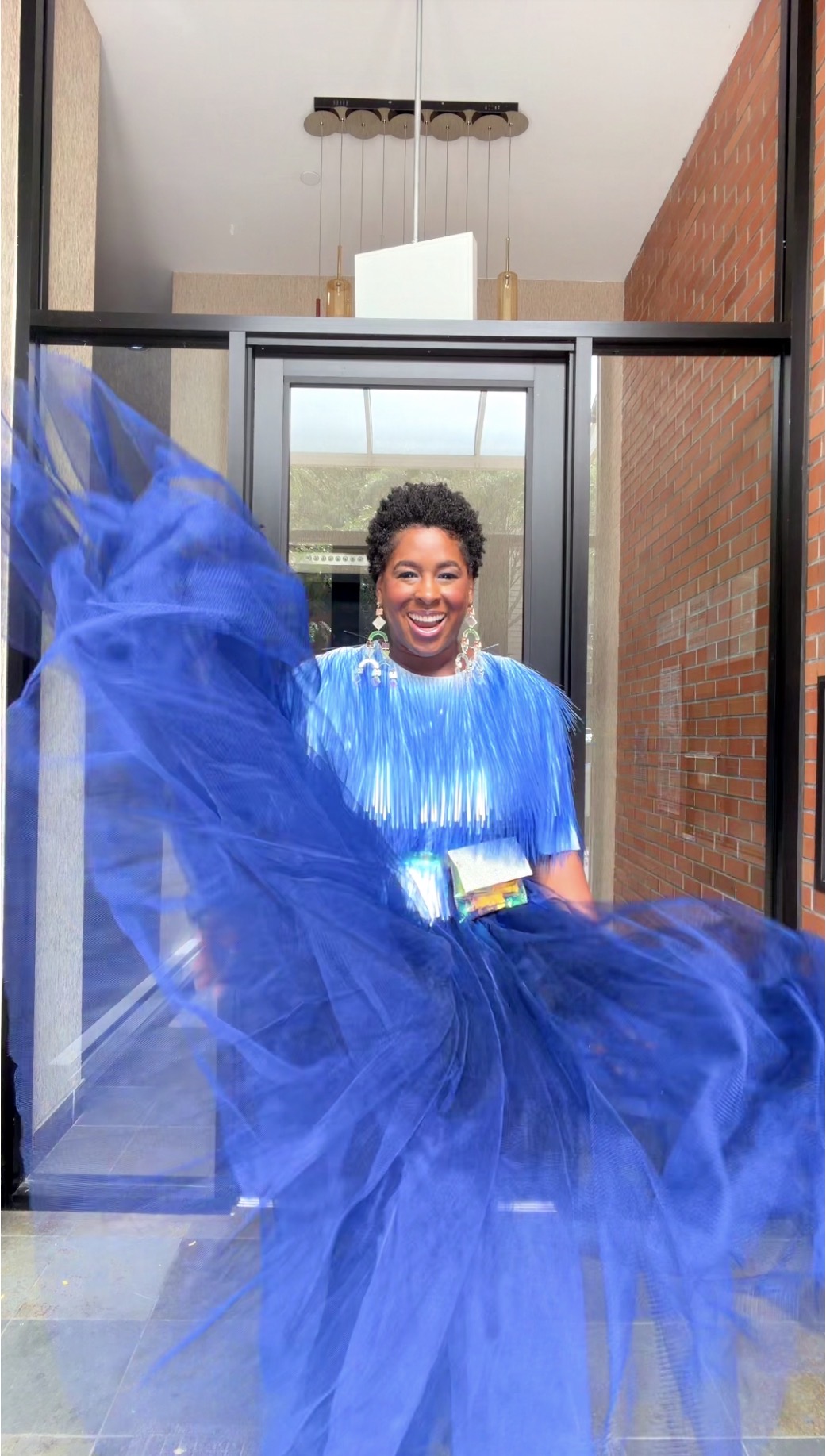 Woman in a blue dress smiling, standing in front of a glass door, with a flowing blue fabric in the foreground.