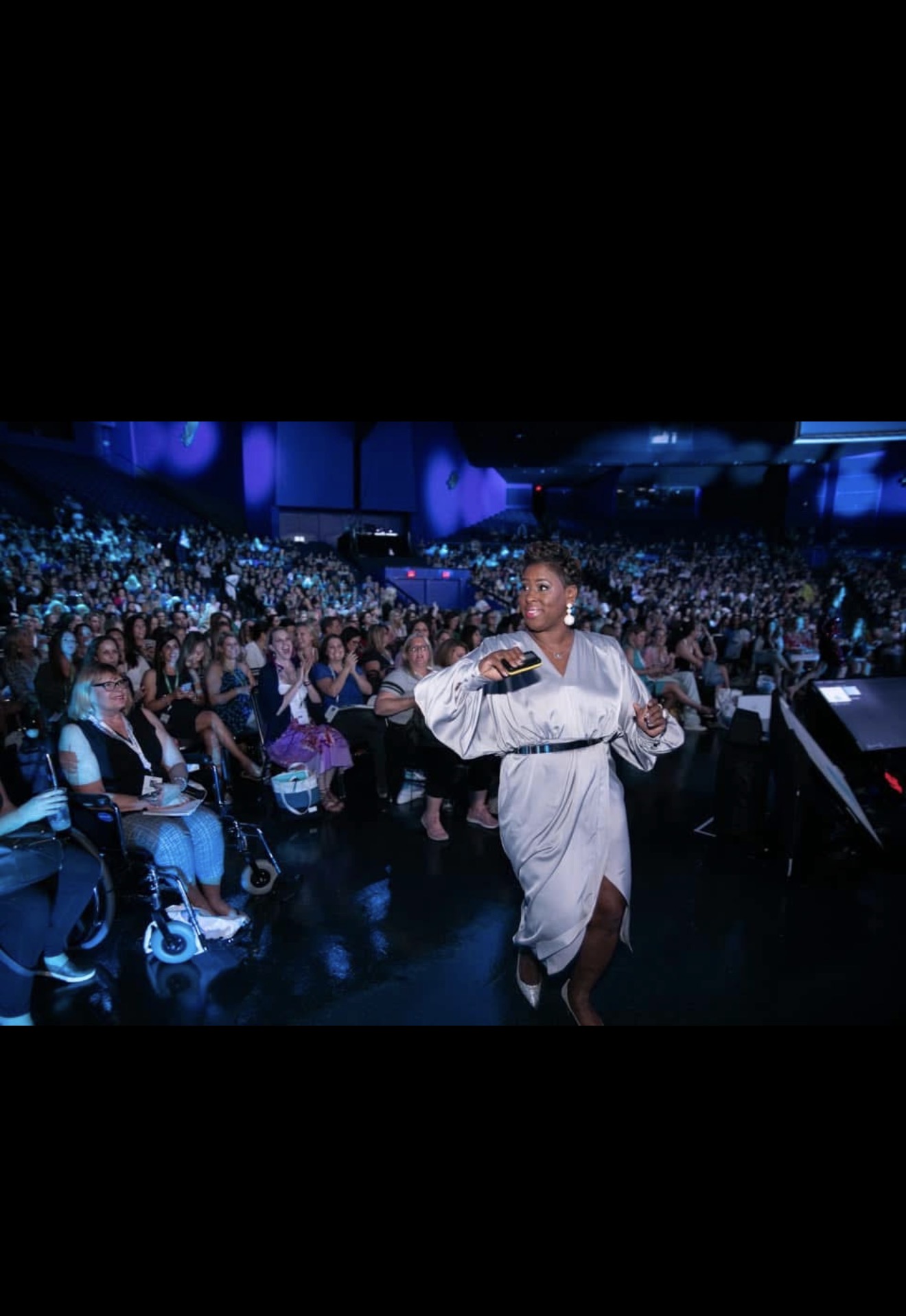 Performer walking on stage in front of large audience with blue lighting, some seated and some standing, in a dark venue.