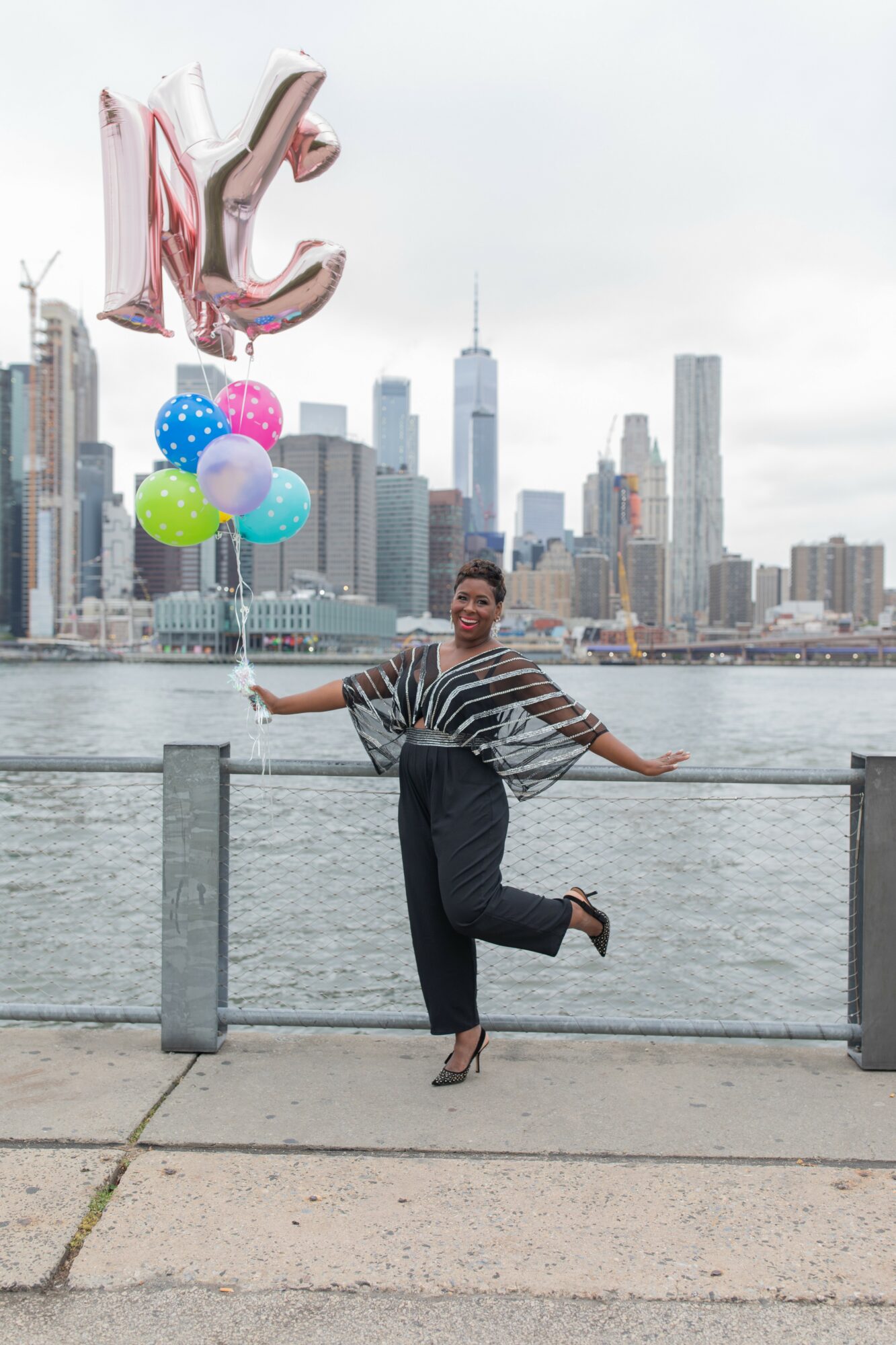 Person standing by water with balloons and a city skyline in the background, smiling and posing with arms outstretched.