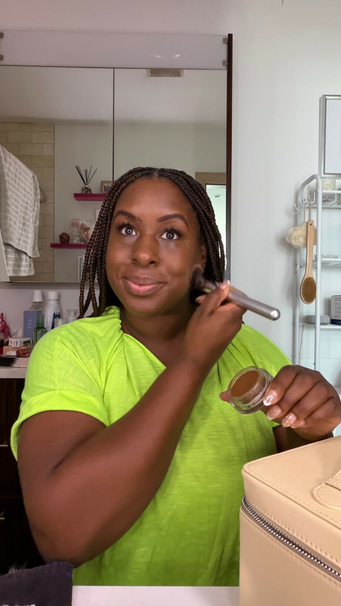Woman applying makeup with a brush in front of a mirror, wearing a bright green shirt.