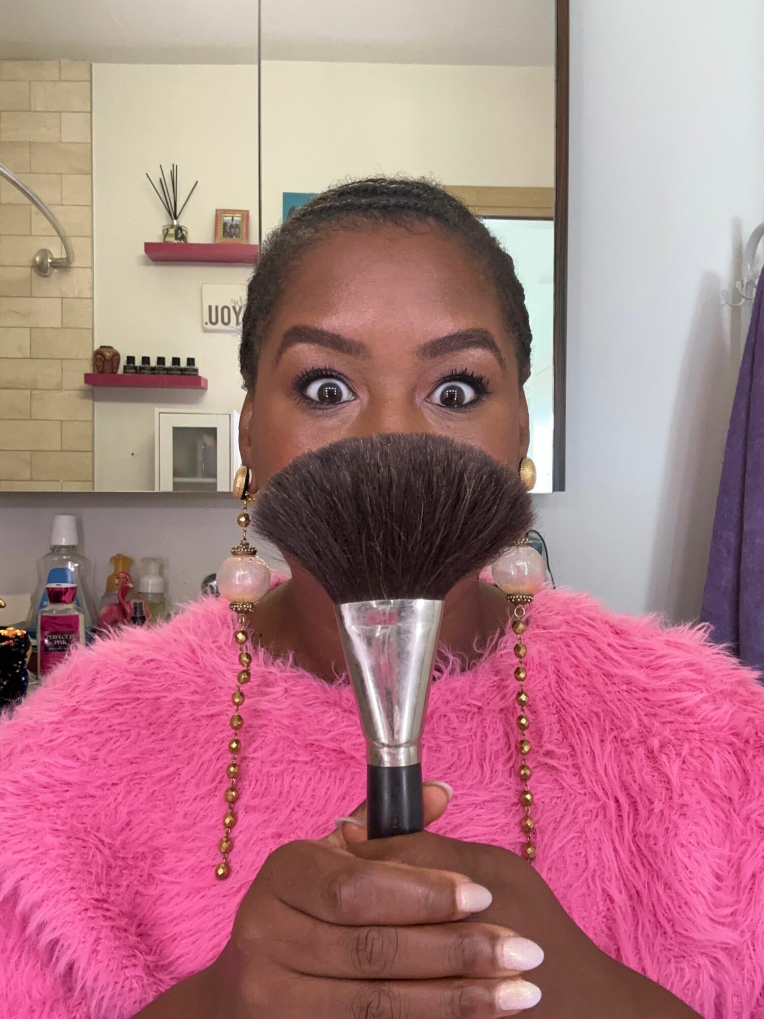 Person in pink fuzzy top holding a makeup brush, with a room background including shelves and a door.