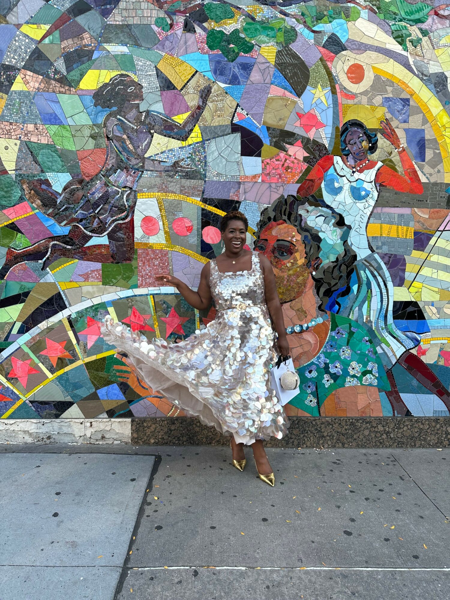 Two women in elegant dresses sit on a colorful mosaic bench, with a vibrant mosaic wall behind them.